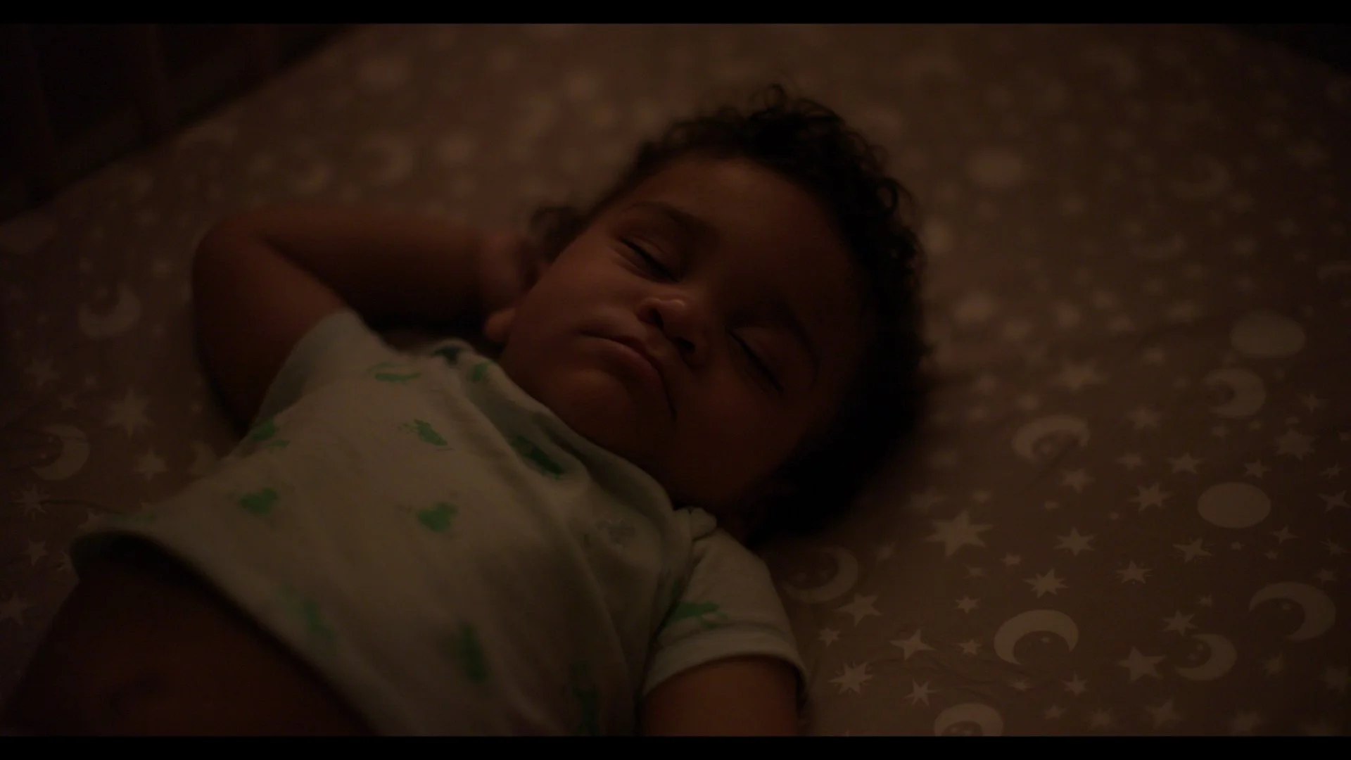 A sleepy baby with curly hair lying on a star and moon patterned bed, resting with one arm behind his head.