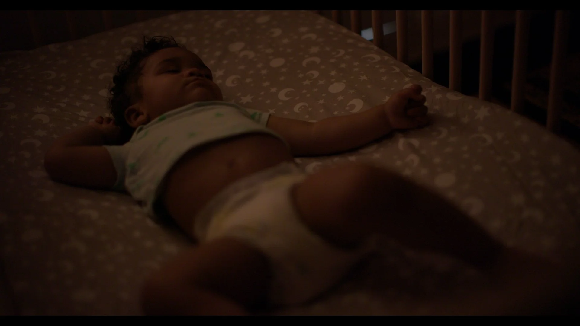 A young child with curly hair sleeping on a bed in a dimly lit room, wearing a white shirt and diaper, lying on their back with one arm stretched out.