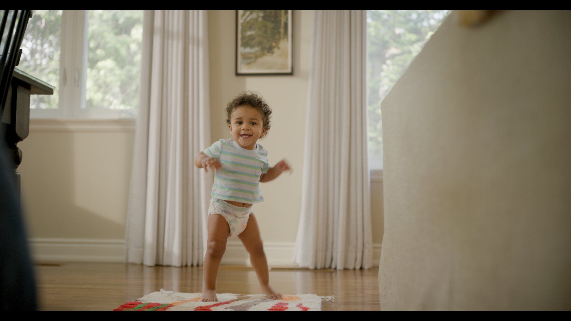 A smiling toddler in a striped shirt and diaper standing on a colorful rug in a living room with curtains and a picture frame on the wall.