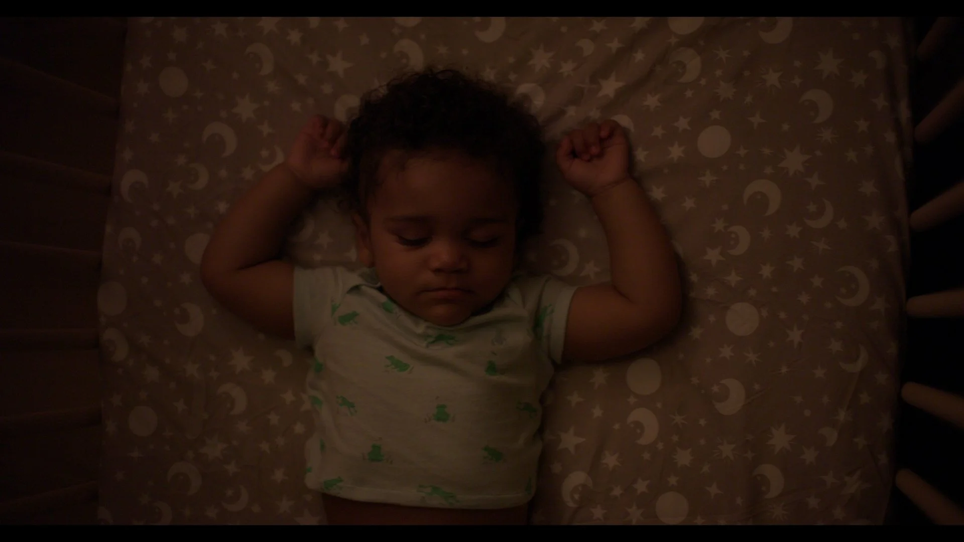 A young child with curly hair sleeping on a bed with a patterned sheet featuring moons, stars, and circles. The child is lying on their back with arms bent and fists near their head, wearing a light-colored shirt with a green pattern.