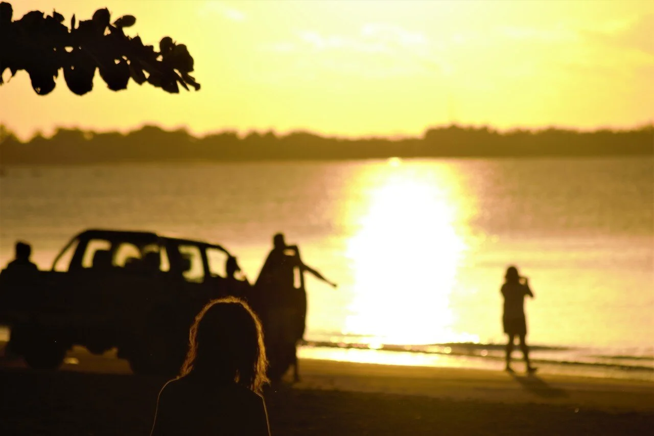 Silhouettes of people near a car by the water during a sunset, with one person taking photos and a woman with curly hair in the foreground.