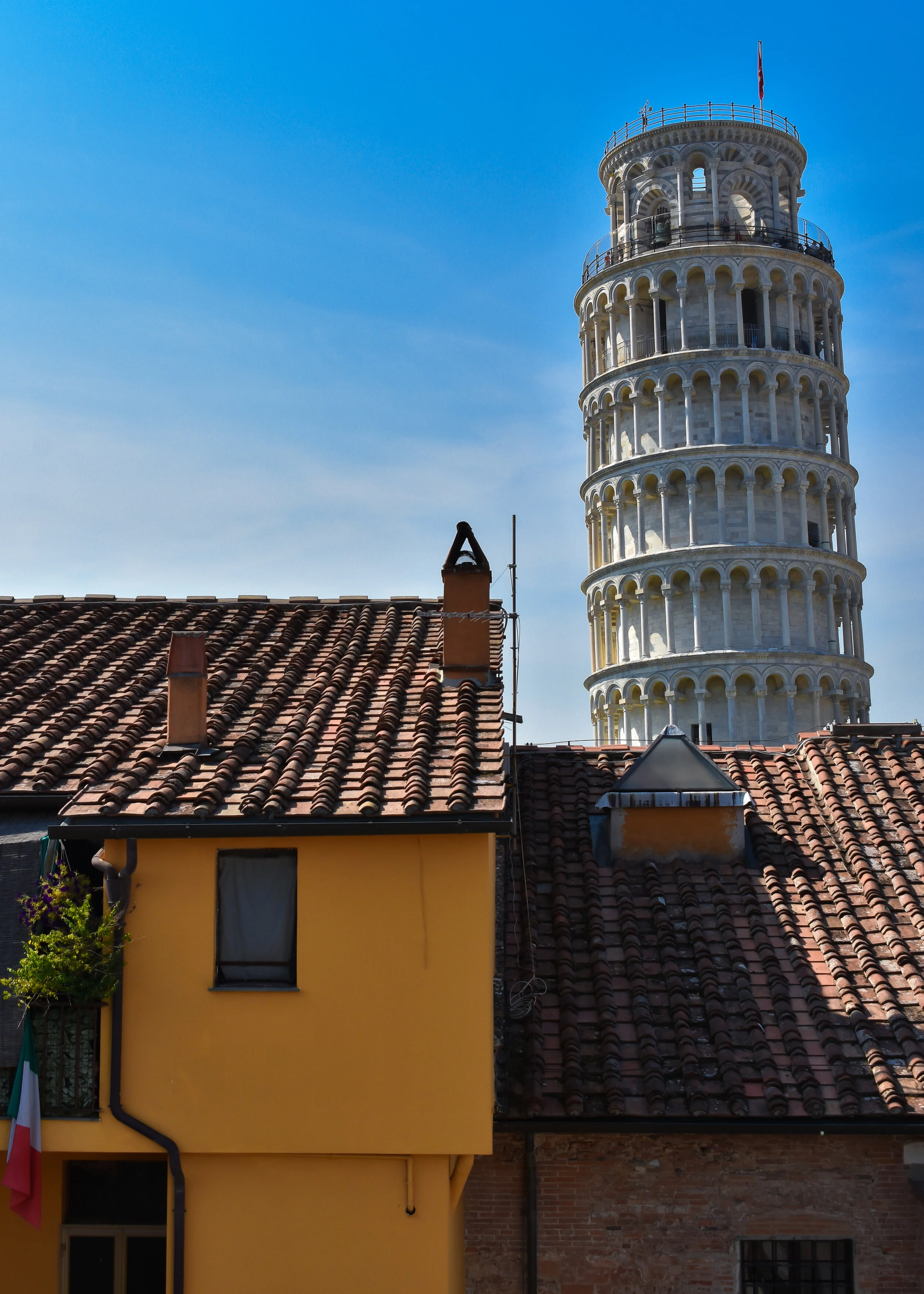 View of a yellow building with a red tiled roof and a chimney, with the Leaning Tower of Pisa in the background against a blue sky.