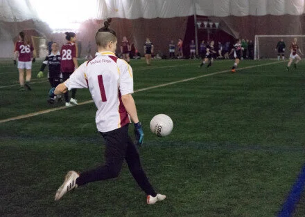 A young boy wearing a white and red sports jersey with the number 1 is walking on an indoor soccer field, with other players and spectators in the background.