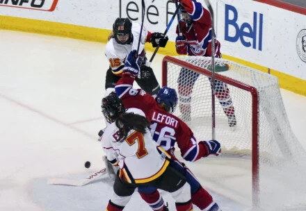Hockey players from two teams battling in front of the goal, with one player attempting to score while the goalie and other players defend.
