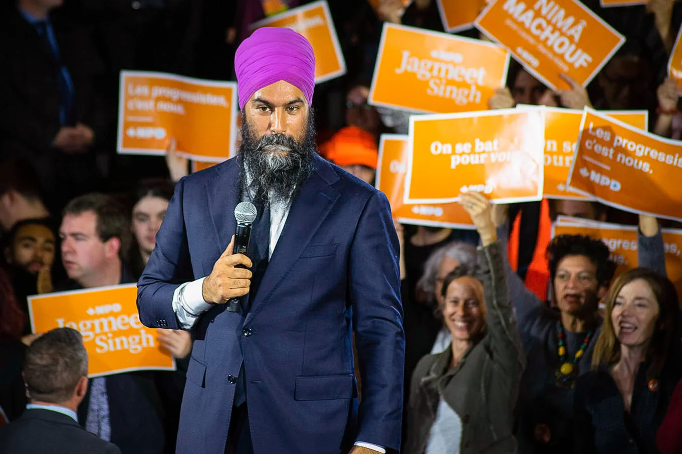A man with a purple turban and beard holding a microphone in front of a crowd holding orange campaign signs.