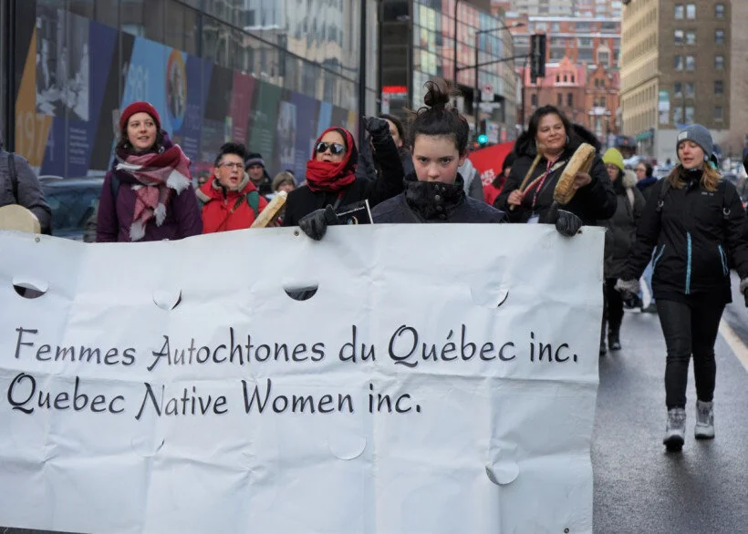 Group of women participating in a protest march on a city street, carrying a banner that reads 'Femmes Autochtones du Québec inc. Quebec Native Women inc.'