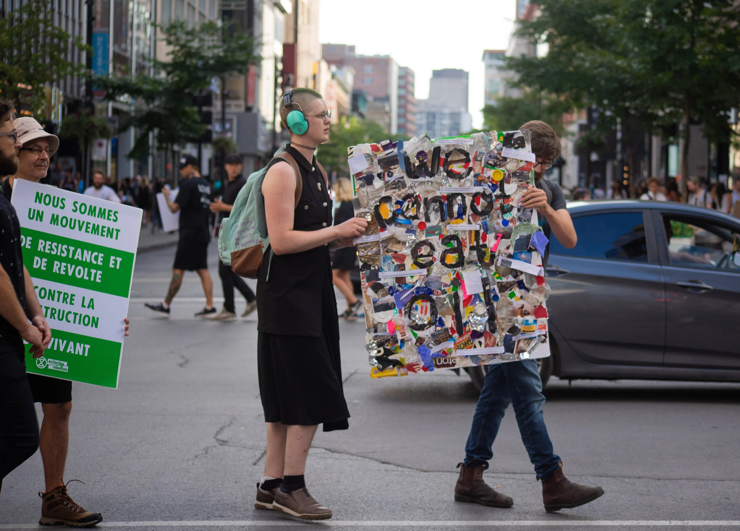 A group of protesters walking down a city street, holding signs advocating for environmental activism and resistance against construction, with one person carrying a collage of various images and materials.