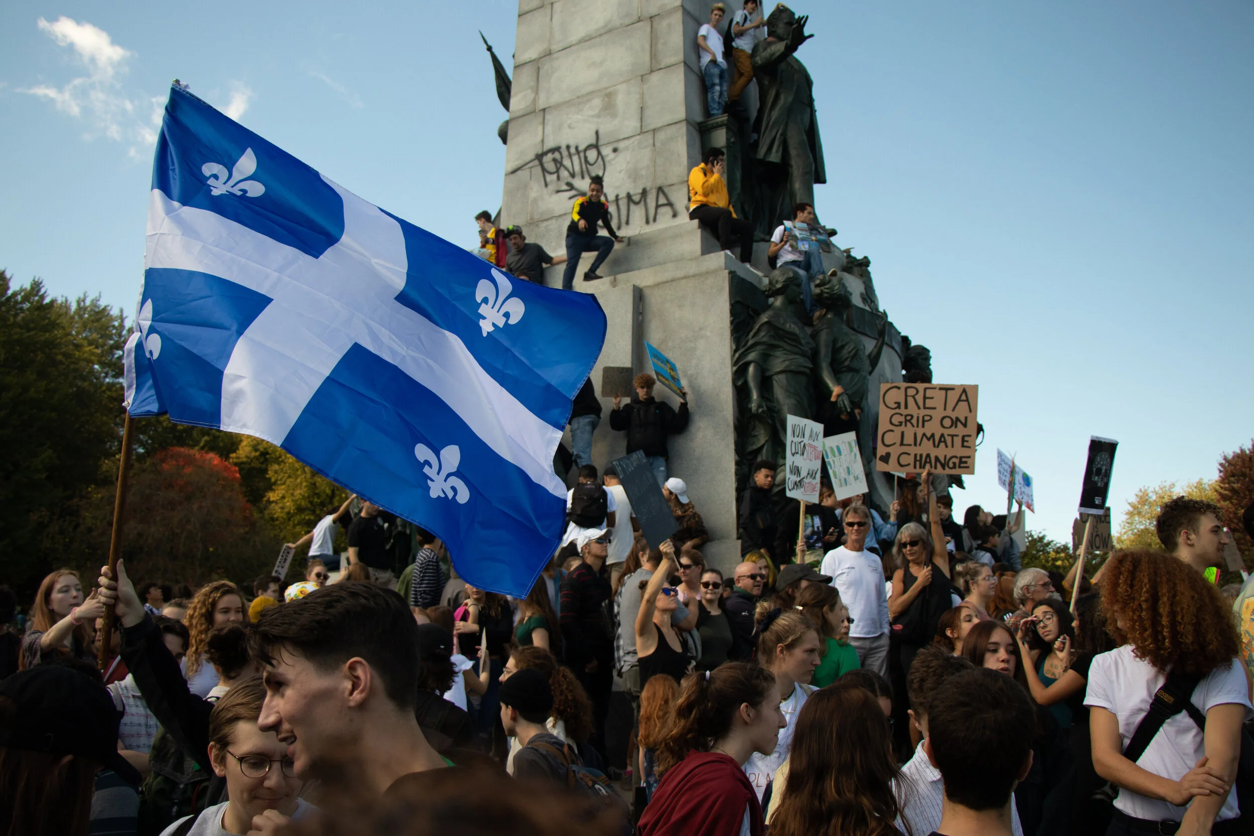Crowd protesting climate change, holding signs that read 'Greta grip on climate change' and 'Non aux changements climatiques,' with people climbing the monument and waving the Quebec flag.