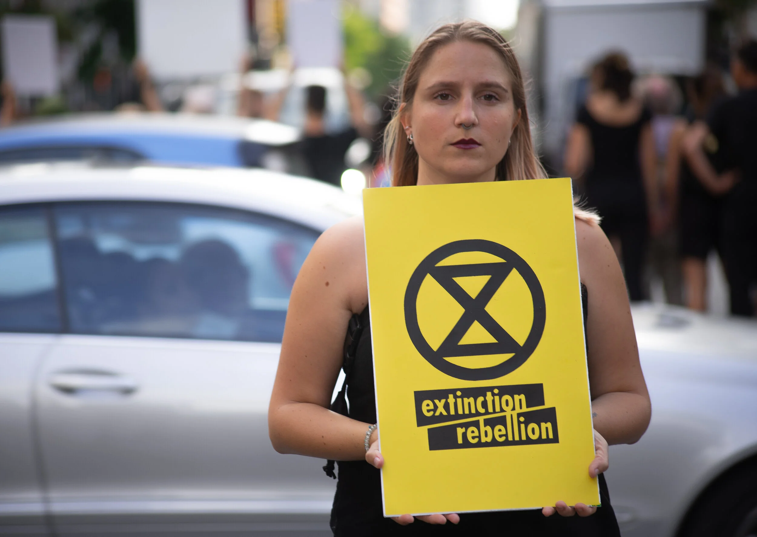 A young woman with platinum blonde hair holding a yellow protest sign with the Extinction Rebellion symbol and text, standing in front of a silver car at a climate protest.