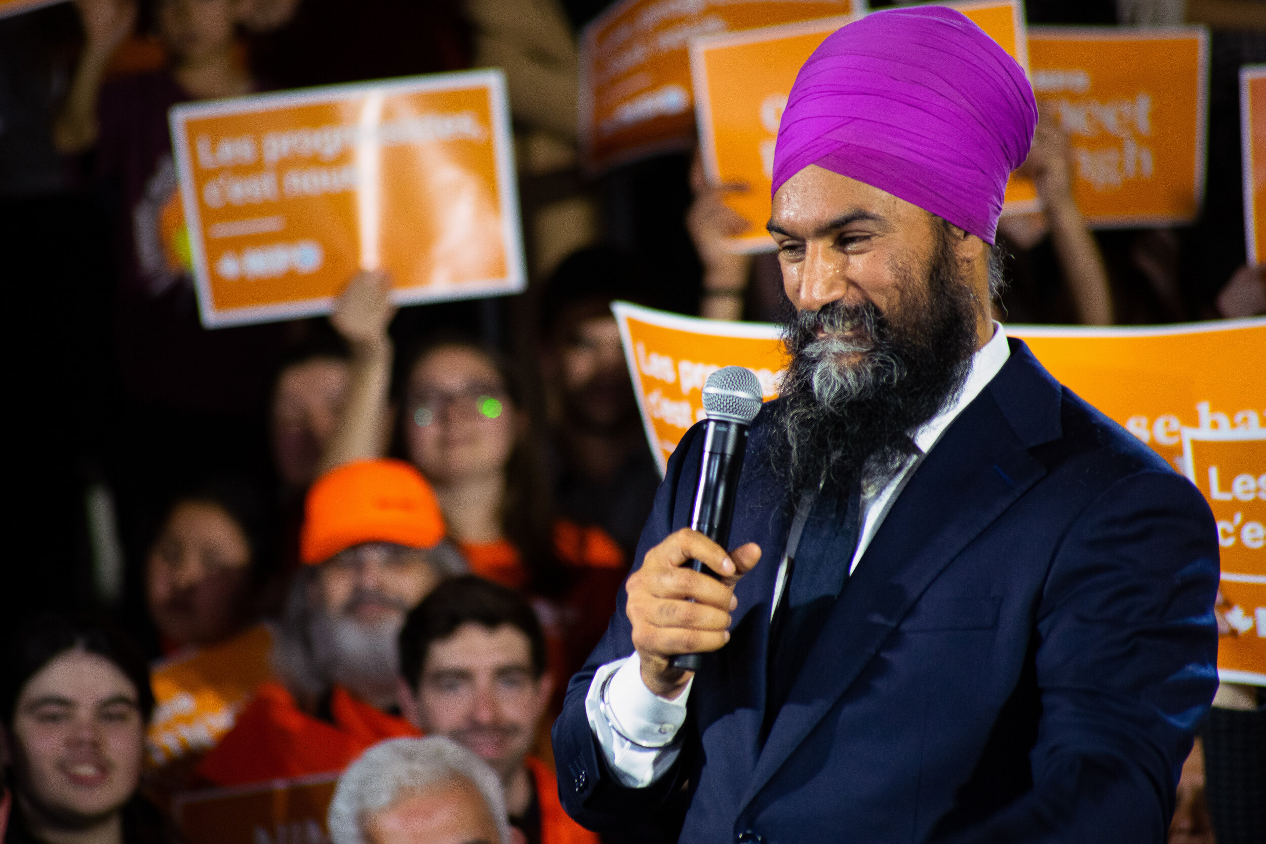 A man with a purple turban and a beard dressed in a dark suit holds a microphone and appears to be speaking at a rally or gathering. Behind him, there are people holding orange signs with white text.
