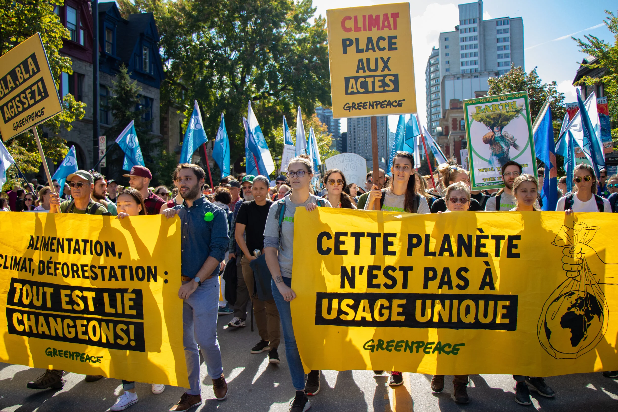 A large group of people participating in a climate change protest march holding signs and banners, including a bright yellow banner with black text in French and English advocating for environmental change, with trees and city buildings in the backgr