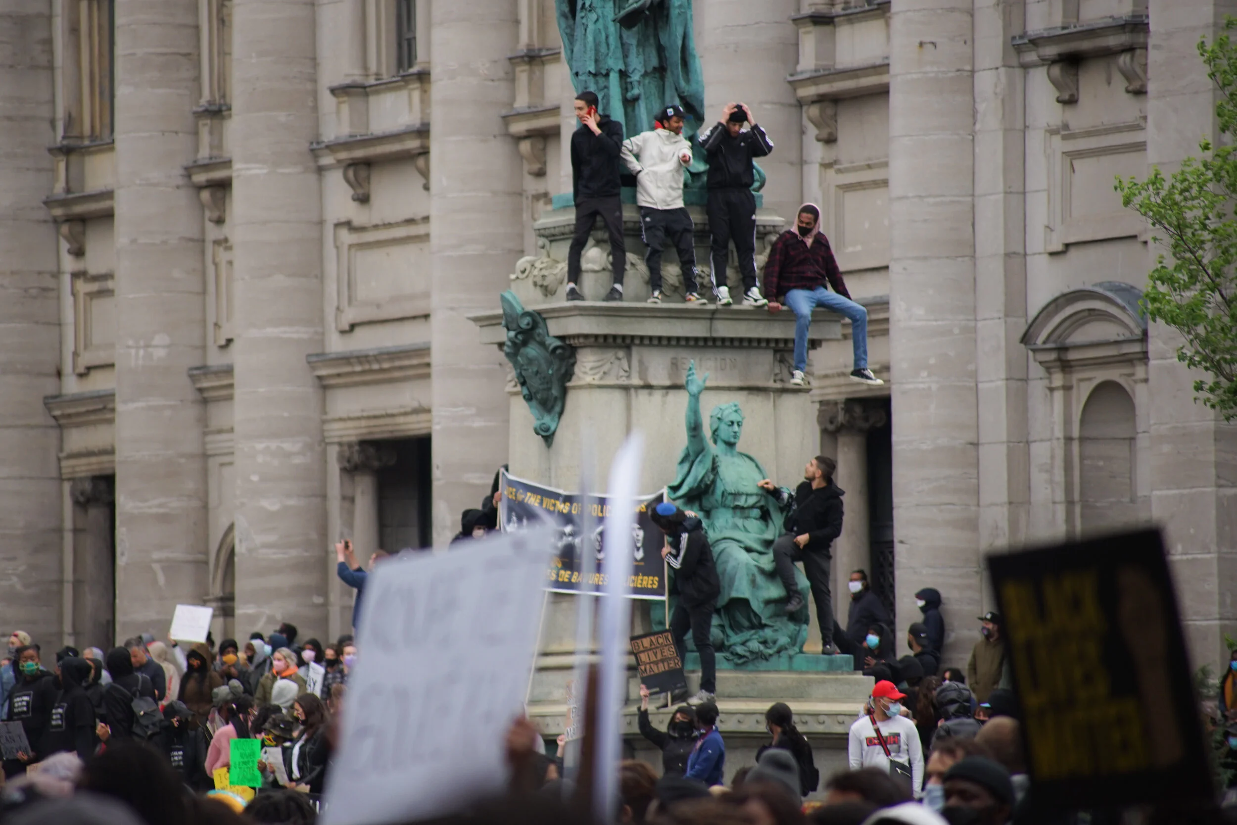 Protesters gathered around a historic building with a statue, some sitting on the statue with one on its lap, and others holding signs, as part of a demonstration.