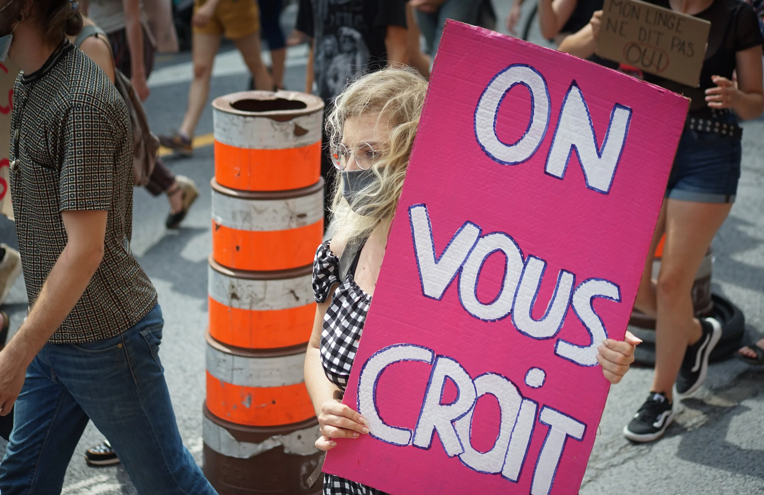 A woman with blonde curly hair and glasses wearing a black face mask holds a pink protest sign that says "ON VOUS CROIT" at a rally. Several people are visible in the background, some holding signs and walking on a street, with orange and white strip