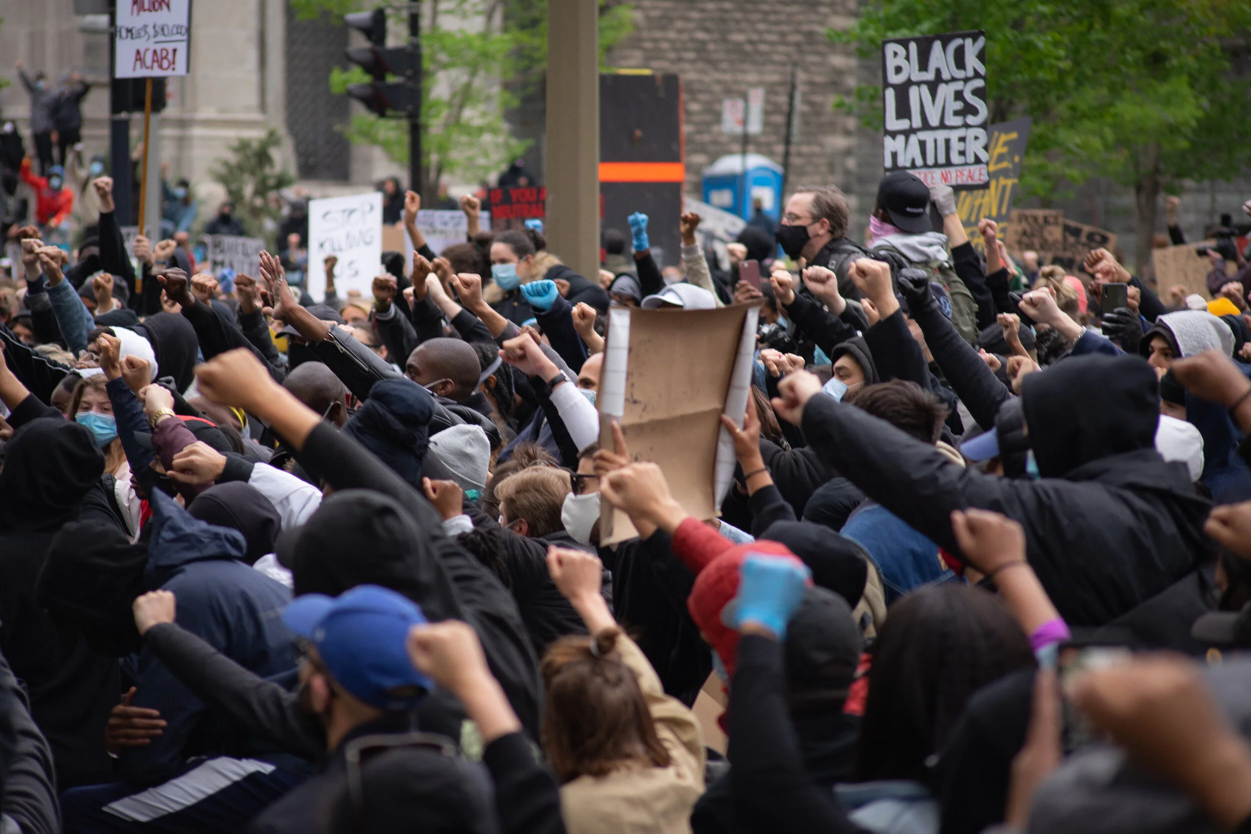 A large crowd of protesters, many wearing face masks, raising their fists in the air during a demonstration for racial justice and Black Lives Matter, with signs like "BLACK LIVES MATTER" and "STOP KILLING" visible in the background.