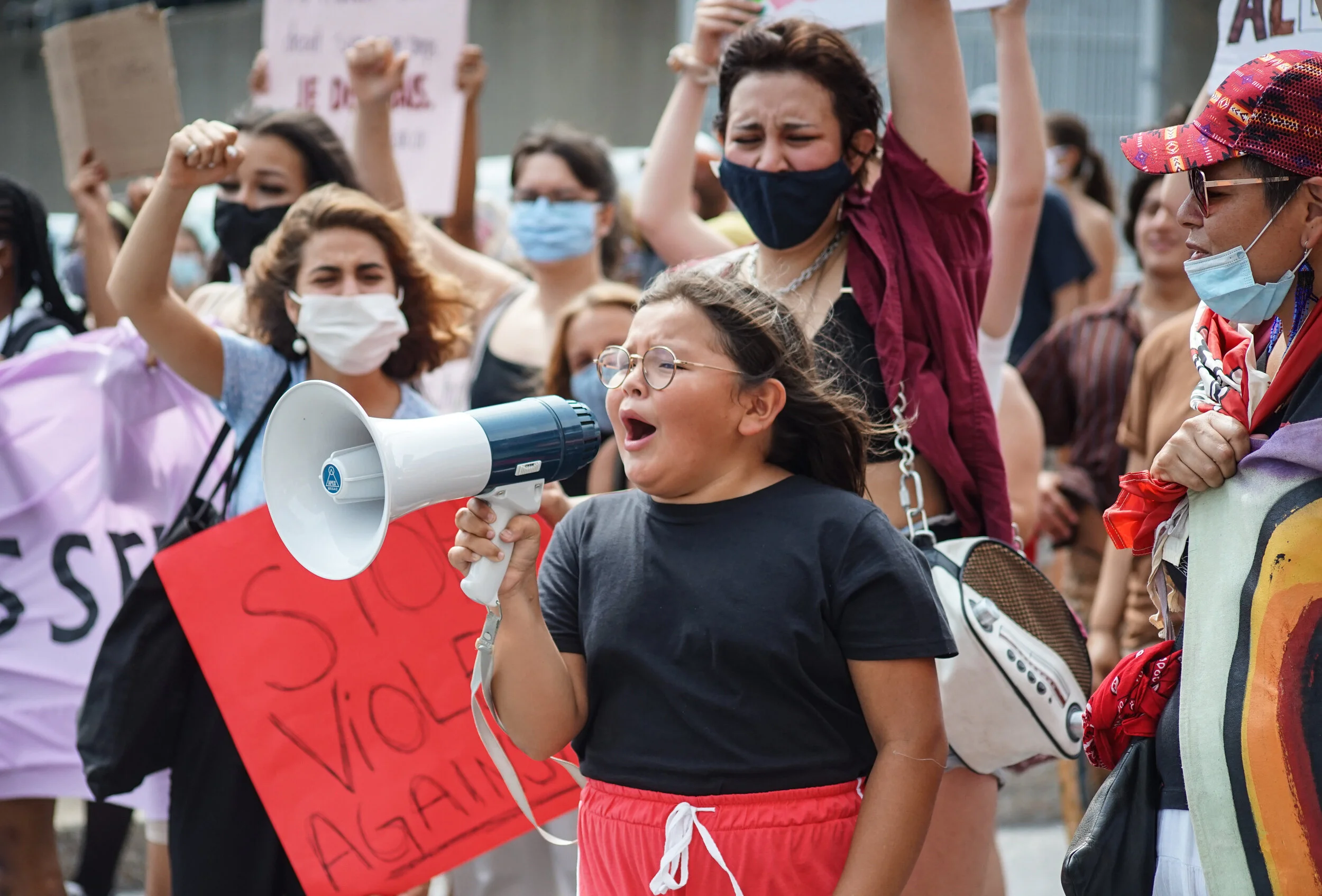 A young girl with glasses and black shirt speaking into a megaphone during a protest, surrounded by women wearing masks and holding signs.