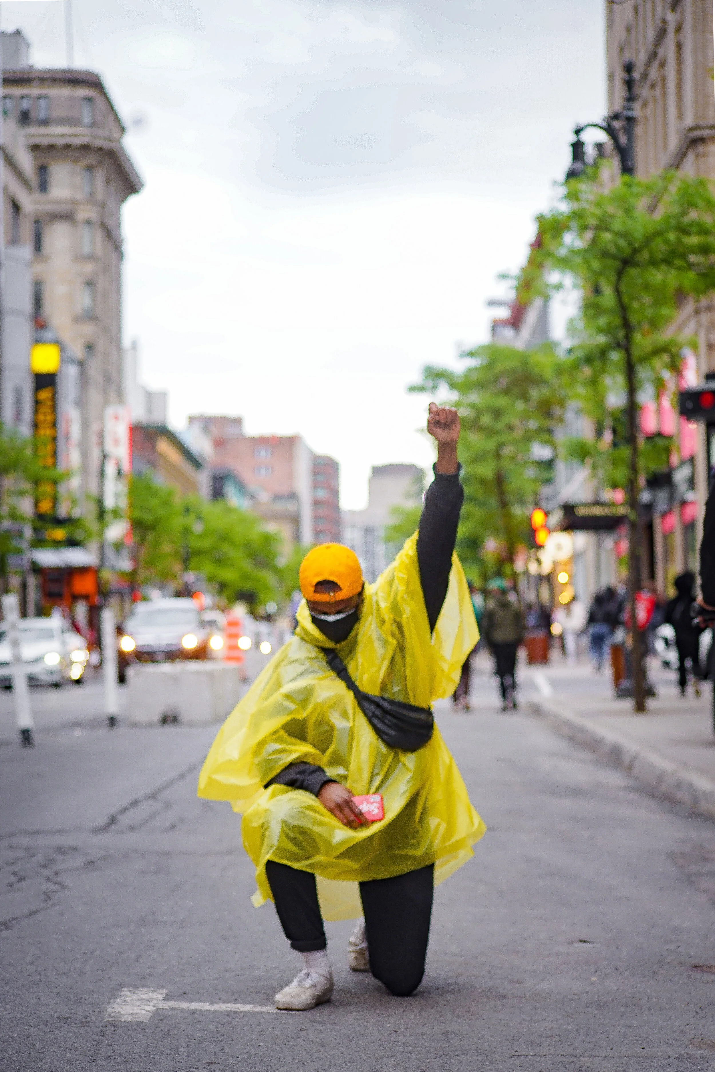 A person wearing a yellow rain poncho, black face mask, orange cap, black backpack, and black pants, kneeling on one knee with one arm raised in a fist on a city street during the daytime.