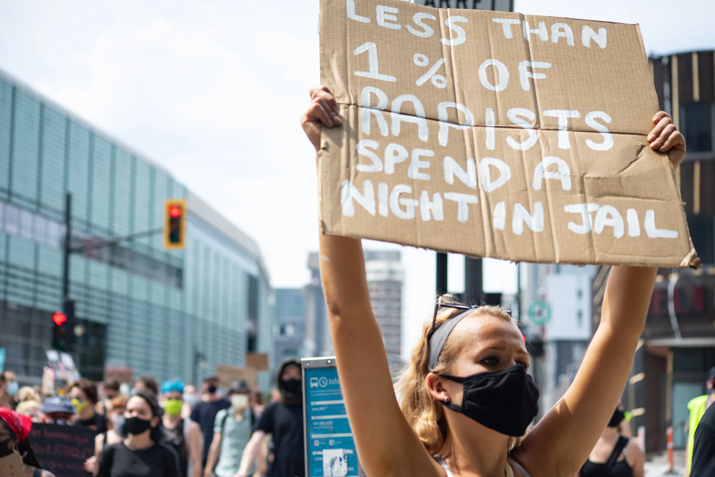 A woman wearing a black face mask holding a cardboard sign that says "Less than 1% of rapists spend a night in jail" during a protest with a crowd in the background.