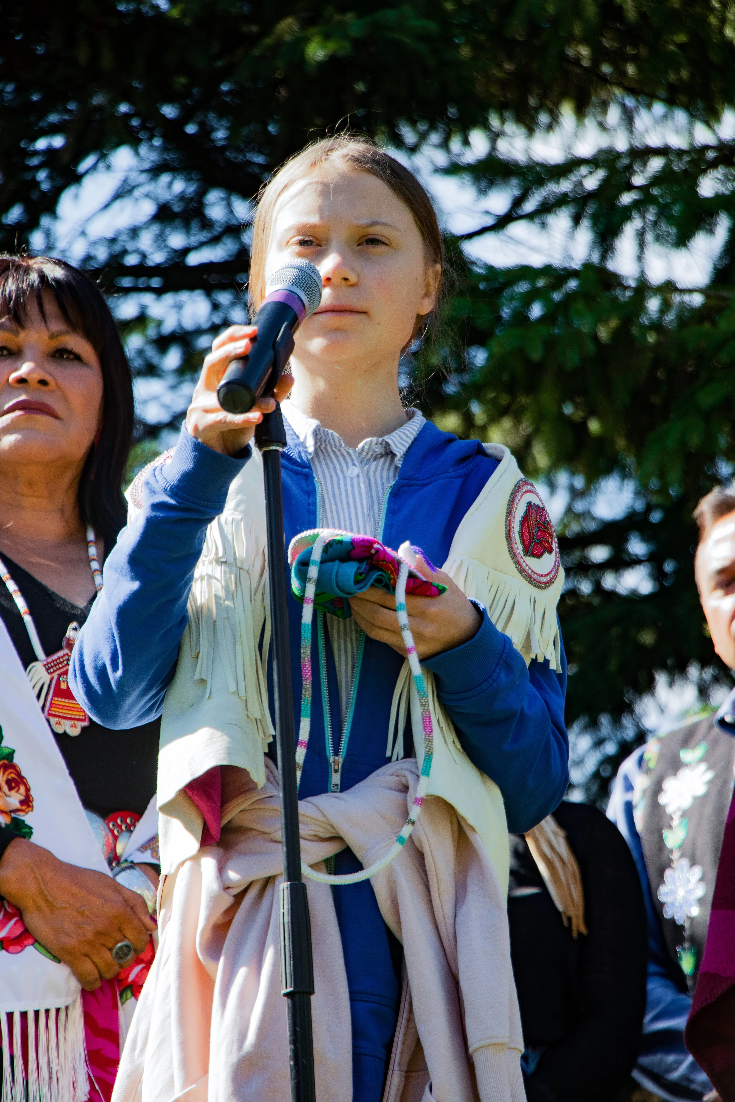 A young woman holding a microphone and speaking at an outdoor event, with other people standing beside her, under trees.