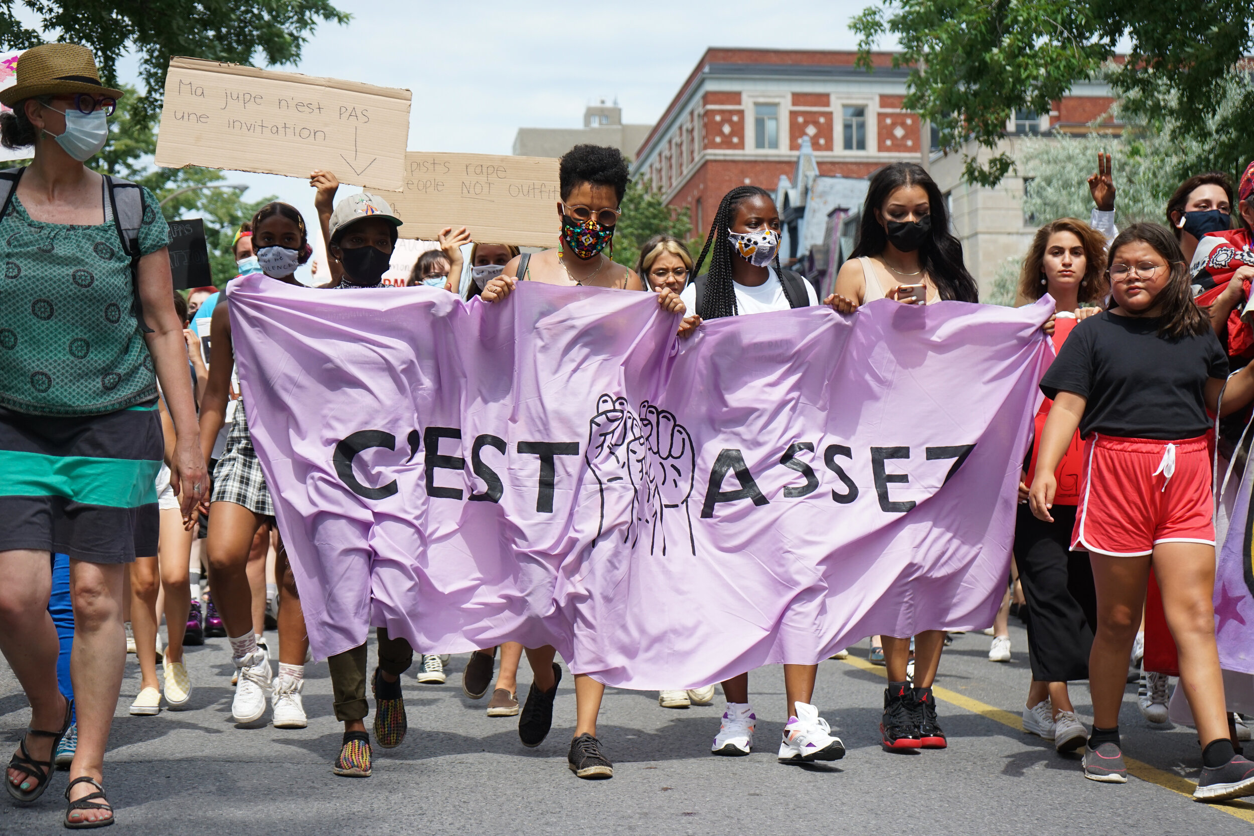 Protesters marching in a street rally, carrying a large purple banner with black text and illustrations, with some holding signs, all wearing masks and casual clothing.