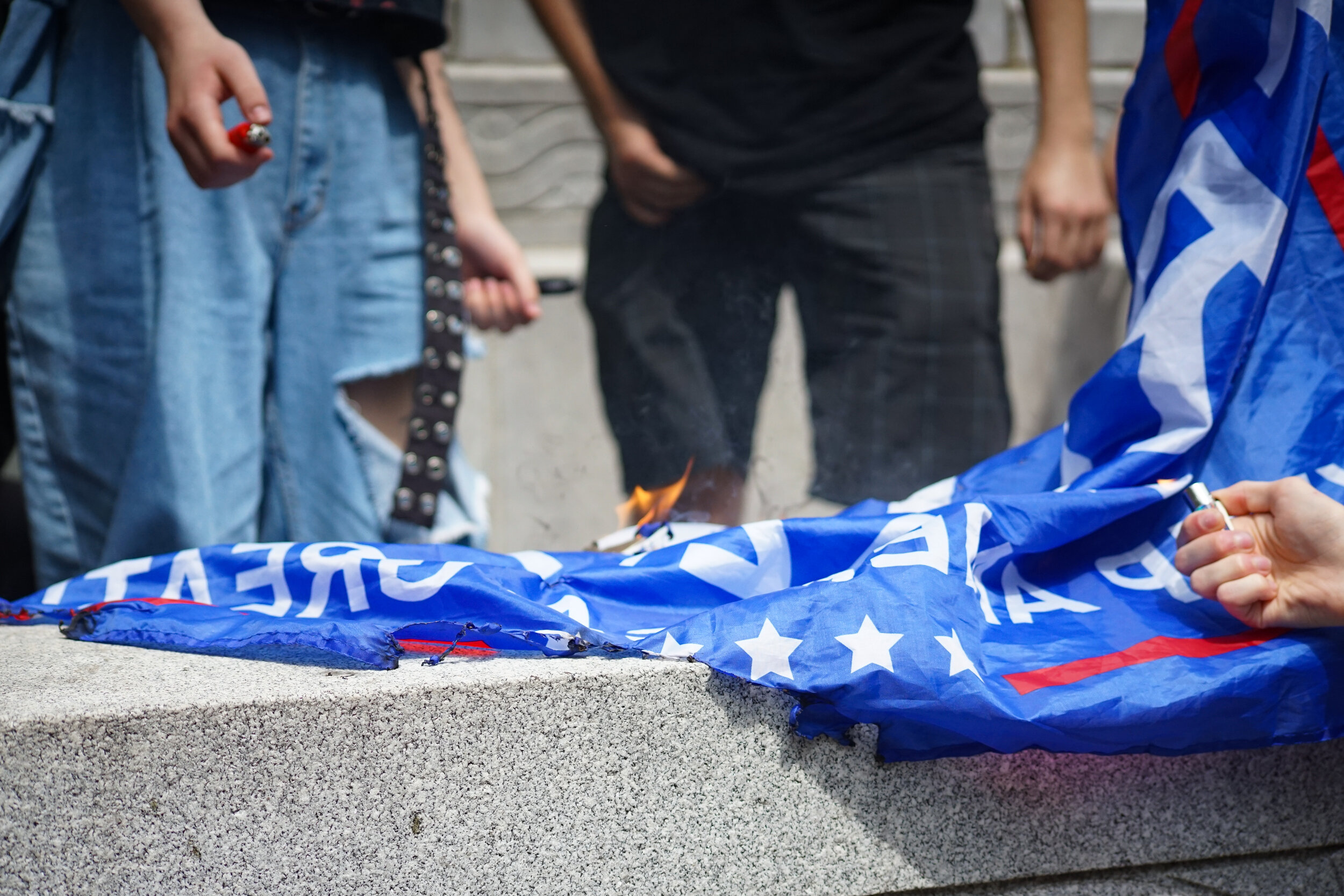 People burning a blue flag with white stars and red and white stripes, likely a political or protest symbol, during a demonstration.
