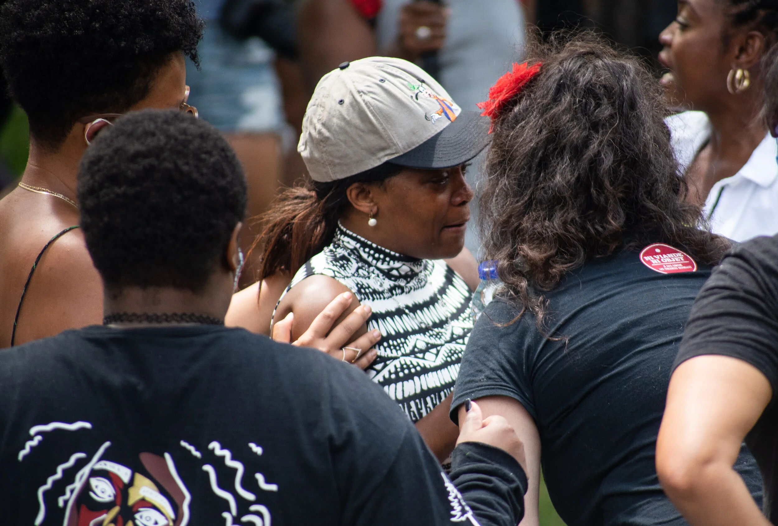 A group of women and girls are seen together, some supporting each other and holding hands, during a gathering or protest. One woman wears a patterned black and white top and a cap. The atmosphere appears emotional and supportive.