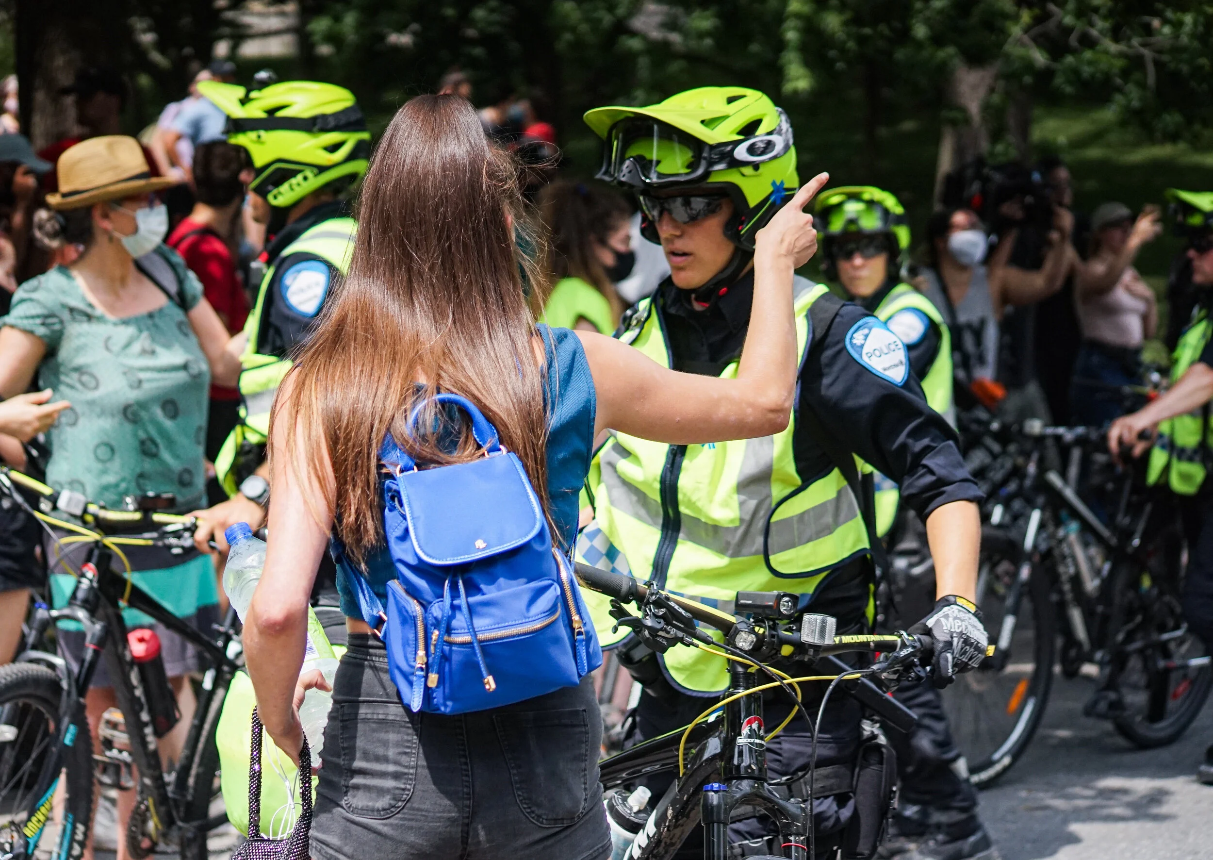A woman with long brown hair talking to a police officer wearing a helmet and yellow vest during a protest or rally with several other people and police officers around.