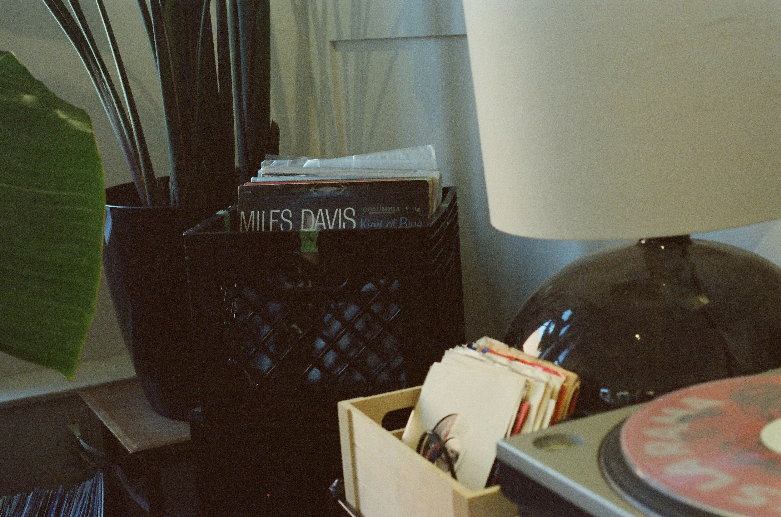 A desk corner with a black wire storage container holding books, a white lamp with a black base, a small wooden box containing papers and miscellaneous items, and a vinyl record player with a record on it.