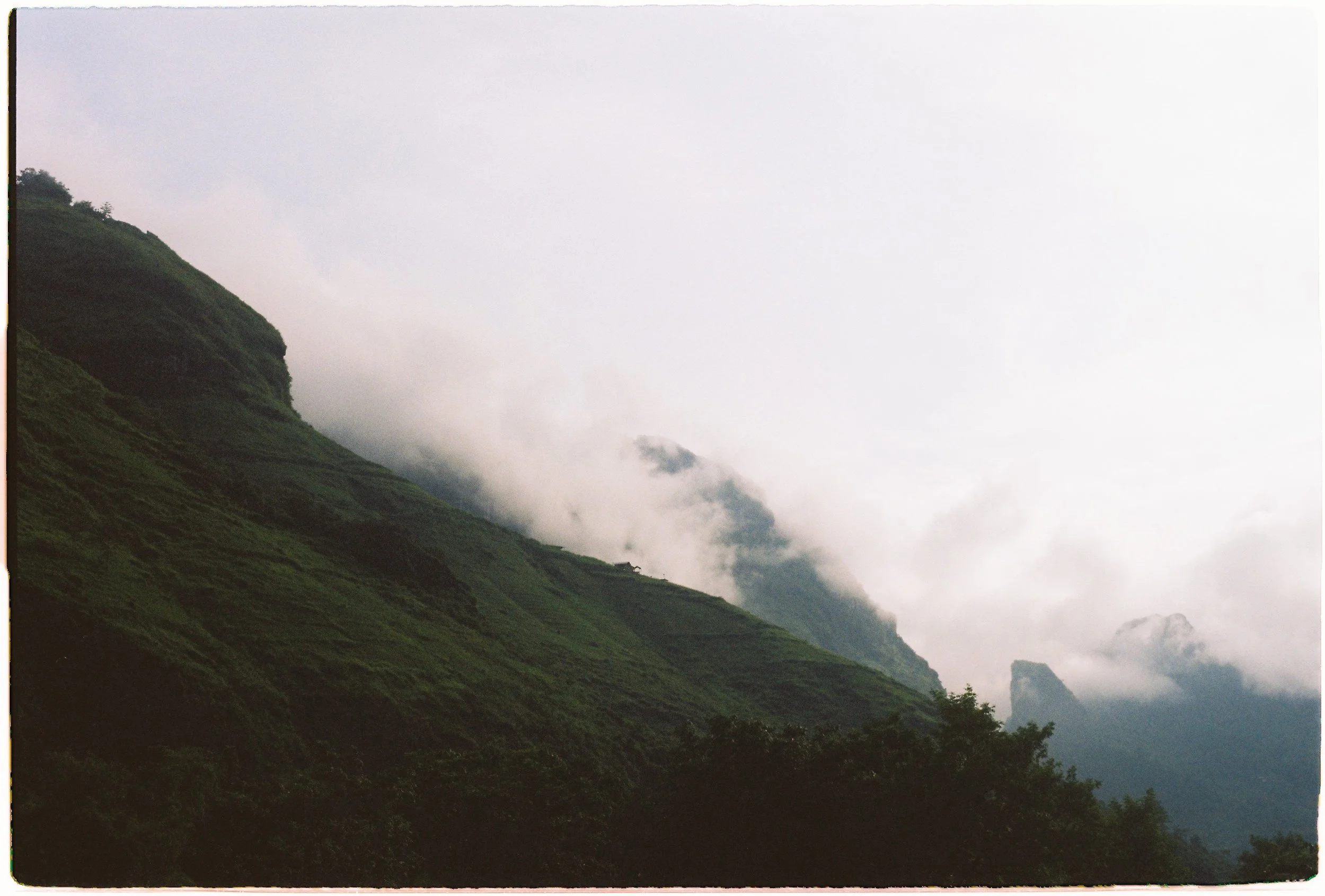 Green mountain covered in mist and clouds with a dark forested area at the base.