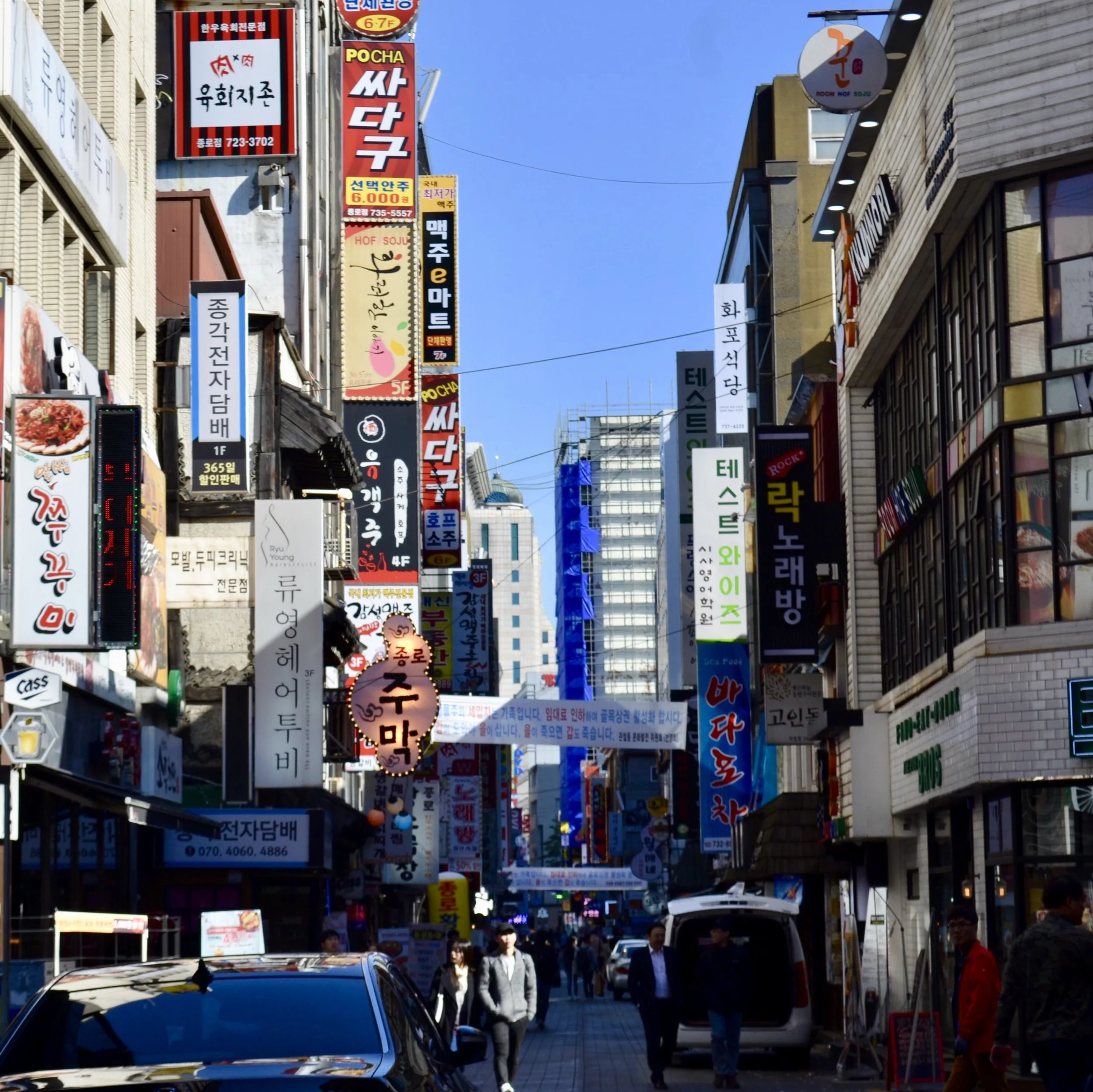 Busy commercial street in Korea with numerous colorful vertical and horizontal signage in Korean, on buildings and above walkways, and pedestrians walking along sidewalks on a sunny day.