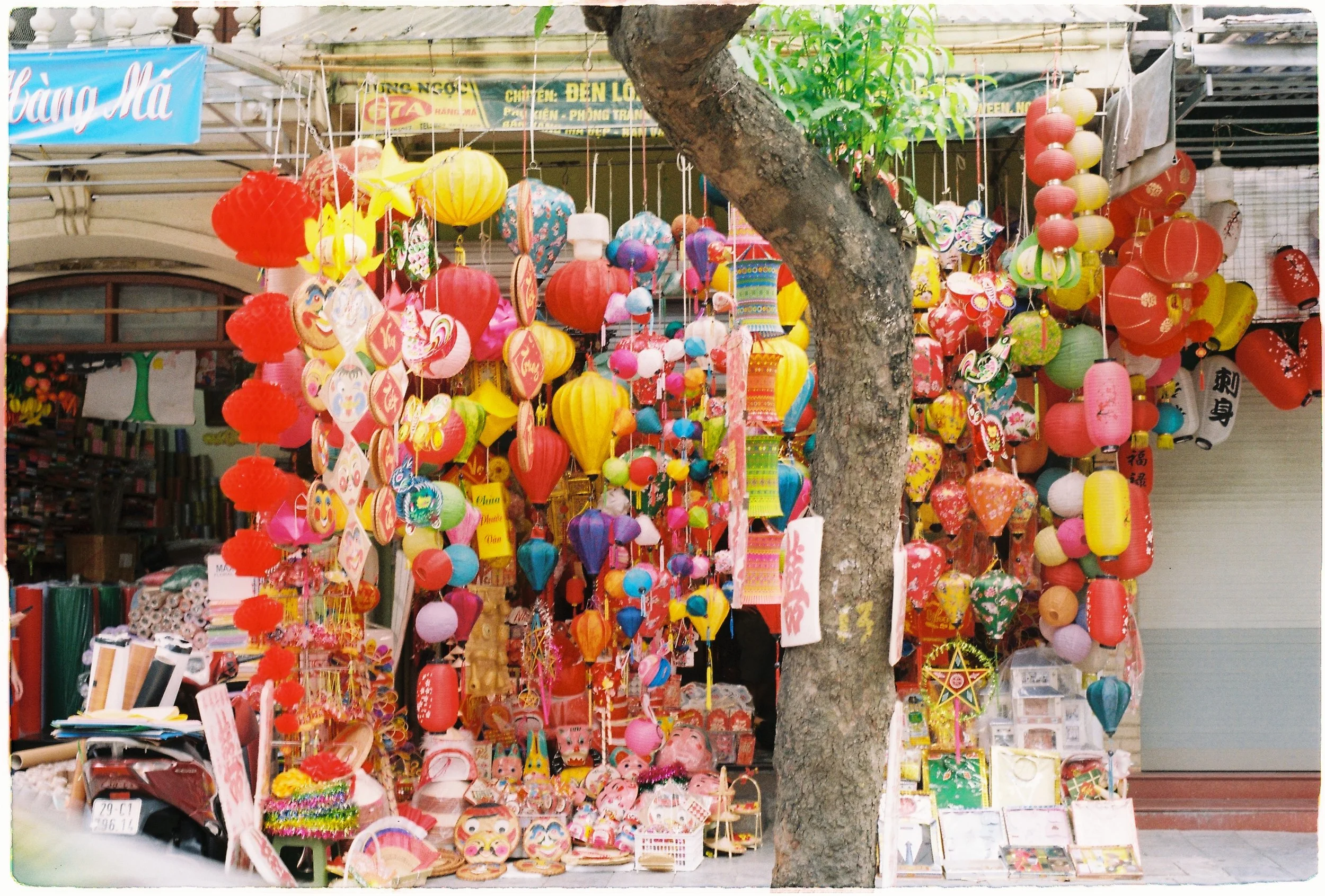 Street vendor stall decorated with colorful paper lanterns, ornaments, and decorations, with a tree in the foreground and shop signs overhead.