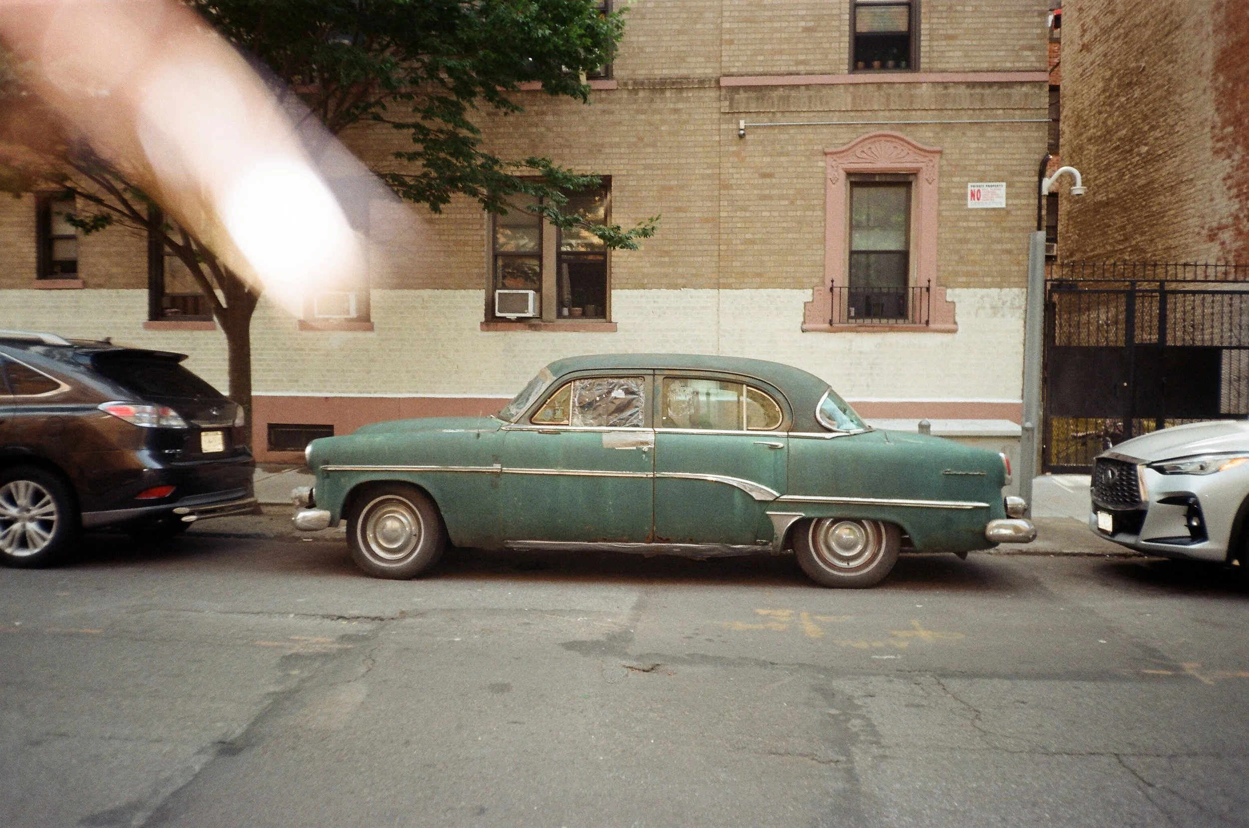An old, weathered green vintage car parked on a city street between modern black and silver cars. The background features a brick building with pink window casings, a tree with green leaves, and a black iron gate.