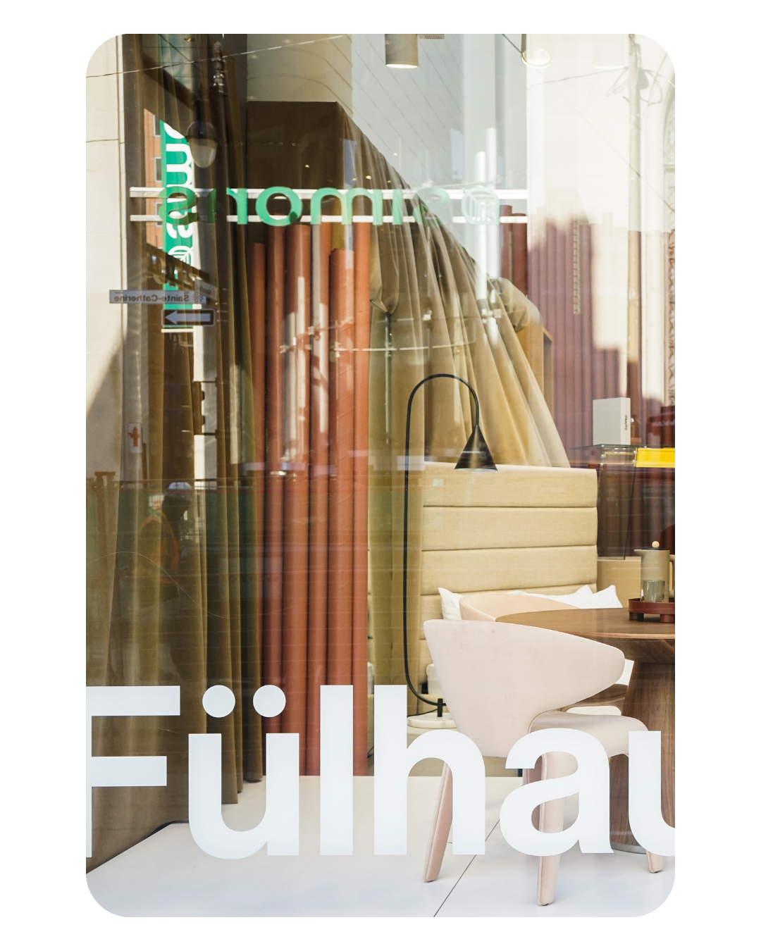 Interior view of a decor store showing fabric rolls, a white chair, a wooden table, and a soft beige headboard. The store window reflects street scenes outside.
