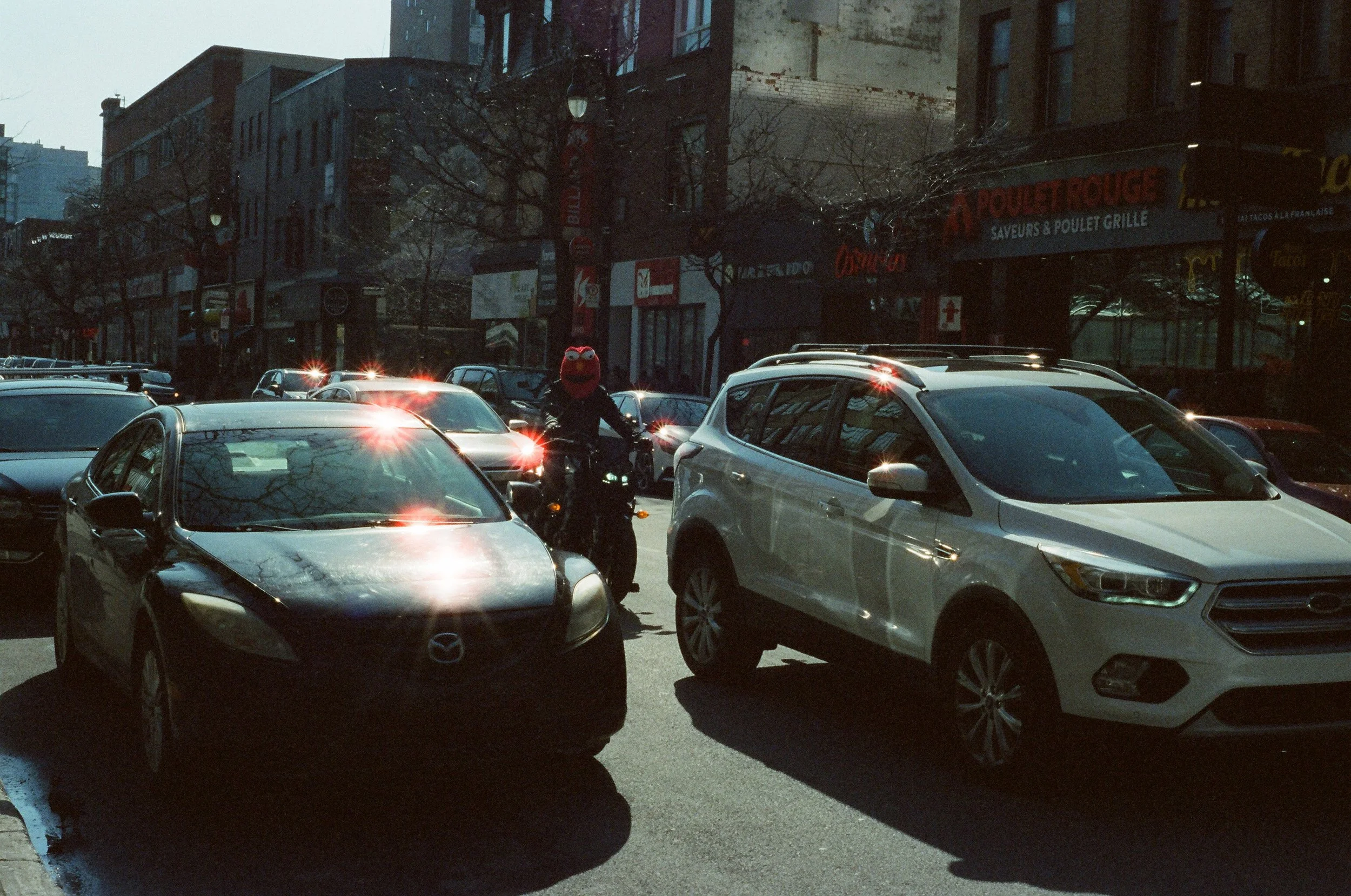 City street scene with parked cars, a person on a bicycle wearing a red helmet, and storefronts on a sunny day.
