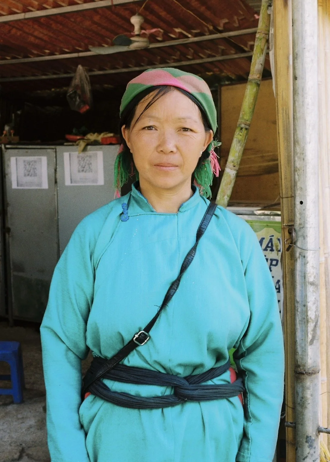 A woman wearing a colorful headscarf and a turquoise traditional outfit standing inside a rustic wooden structure.