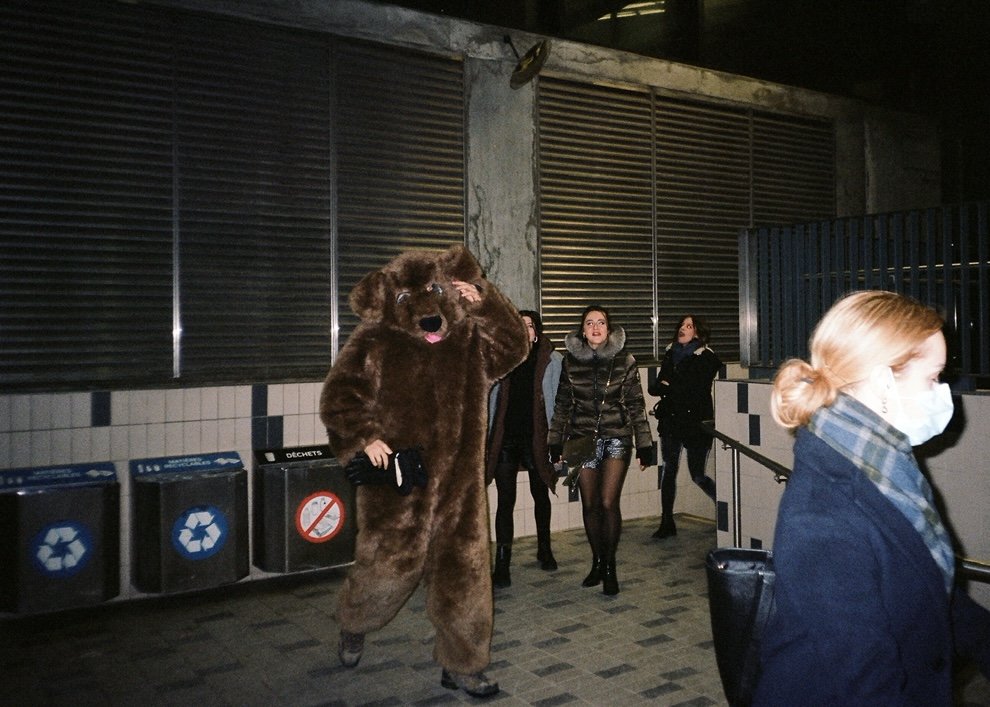 Person dressed in a bear costume walking in an underground station with other people, some wearing masks.