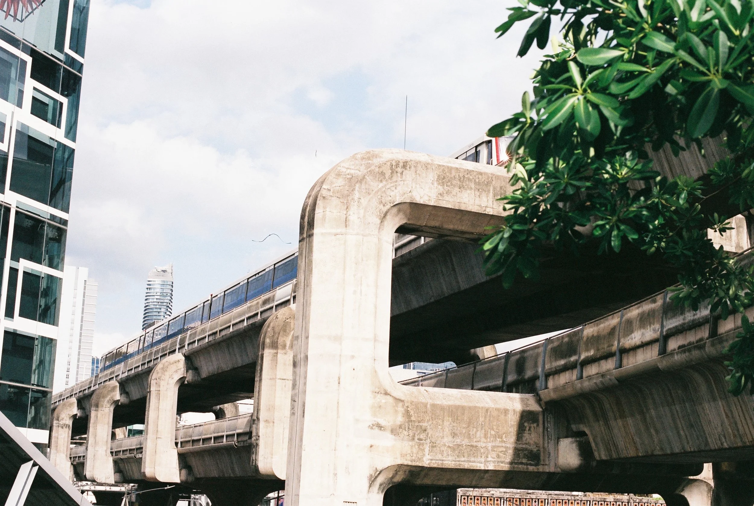 Elevated concrete train tracks with modern buildings and green trees in an urban setting.