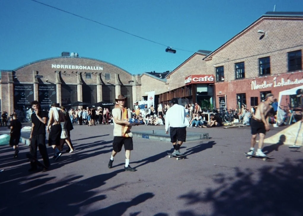 People walking and skateboarding outside Norrebrohallen building in Copenhagen on a sunny day, some sitting near storefronts including a cafe.