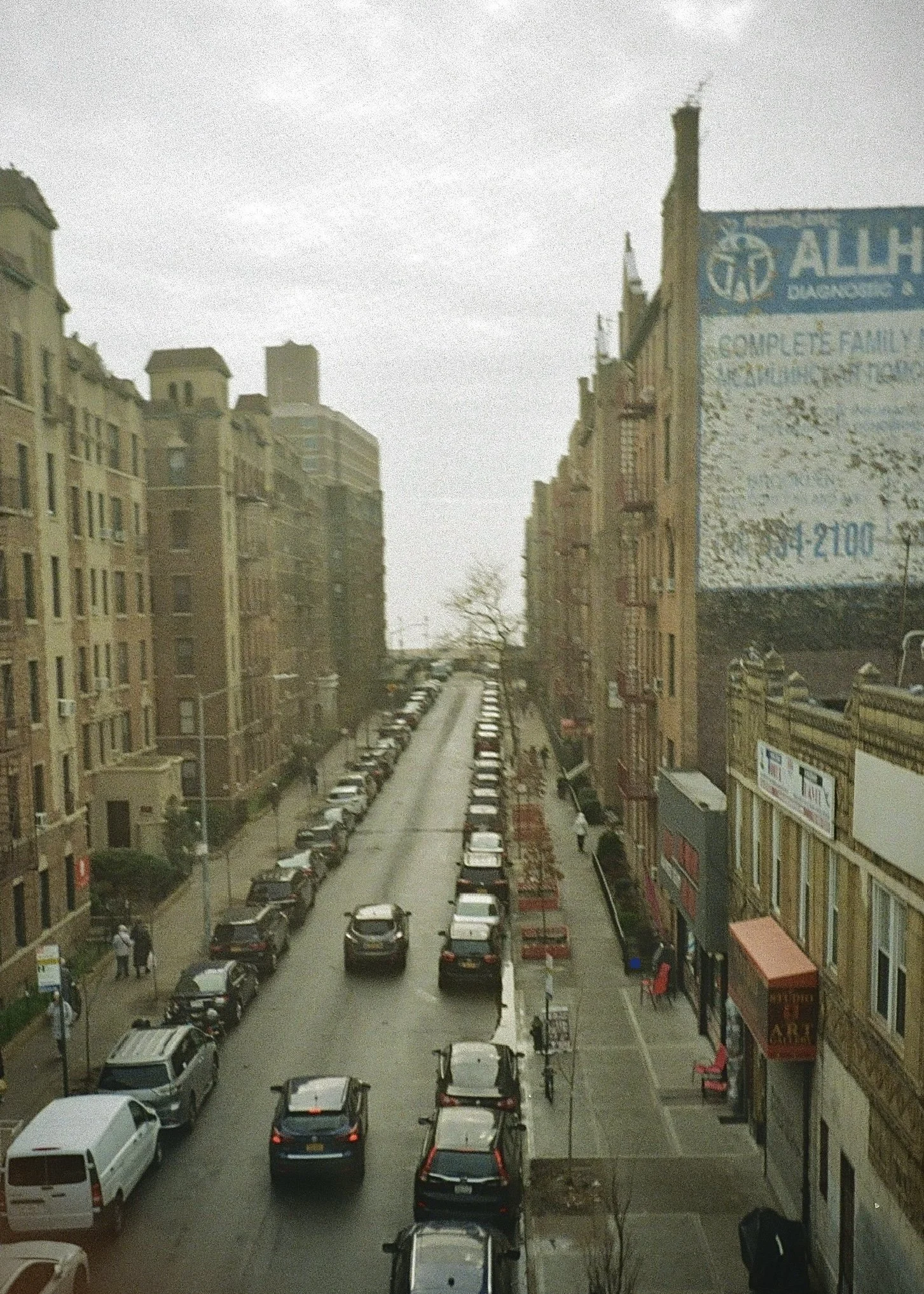 City street view showing parked cars along the curb, multi-story buildings on both sides, and a cloudy sky overhead.