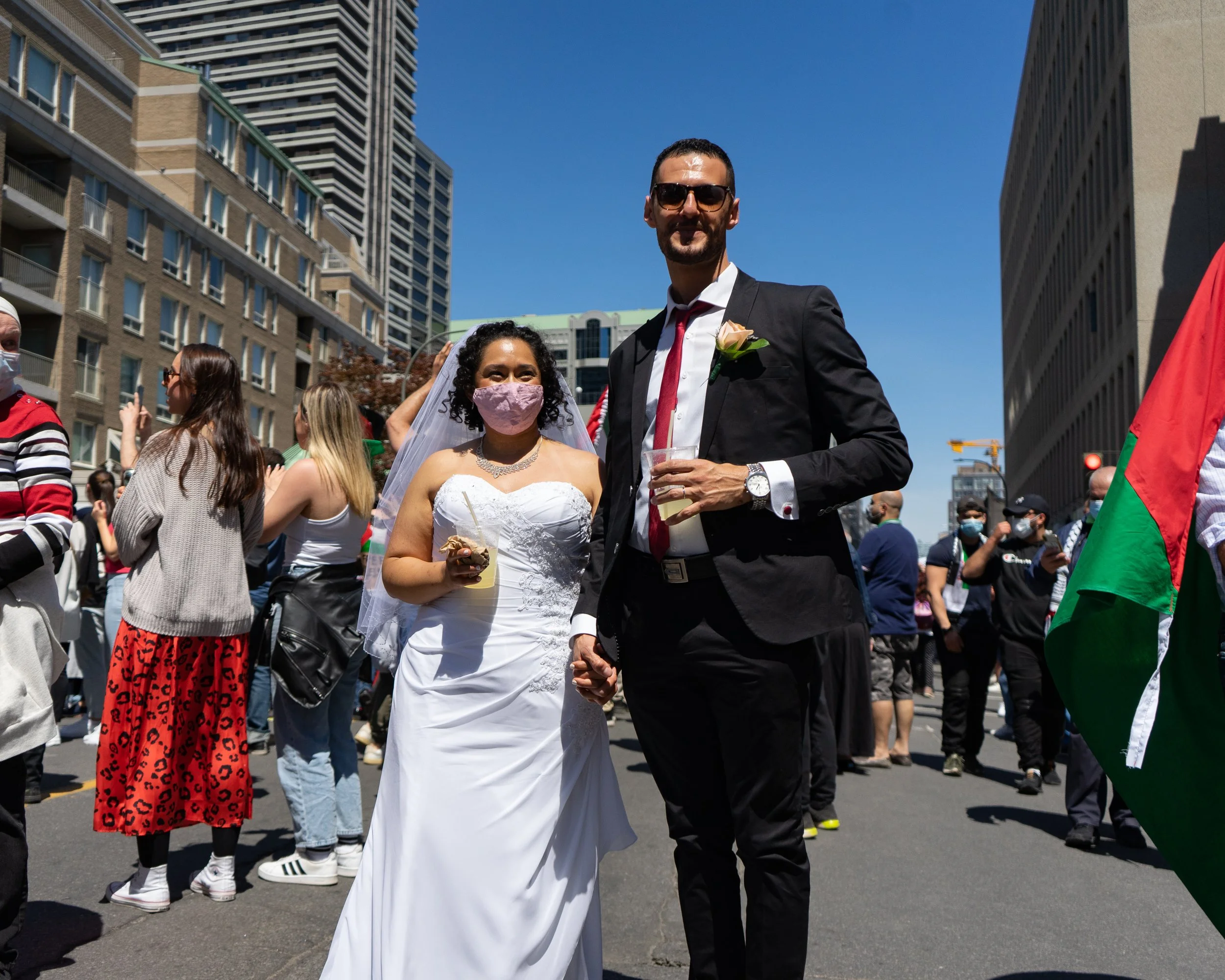 A bride and groom holding hands at an outdoor wedding celebration with people in the background, some wearing masks, in a city street with tall buildings and a clear blue sky.