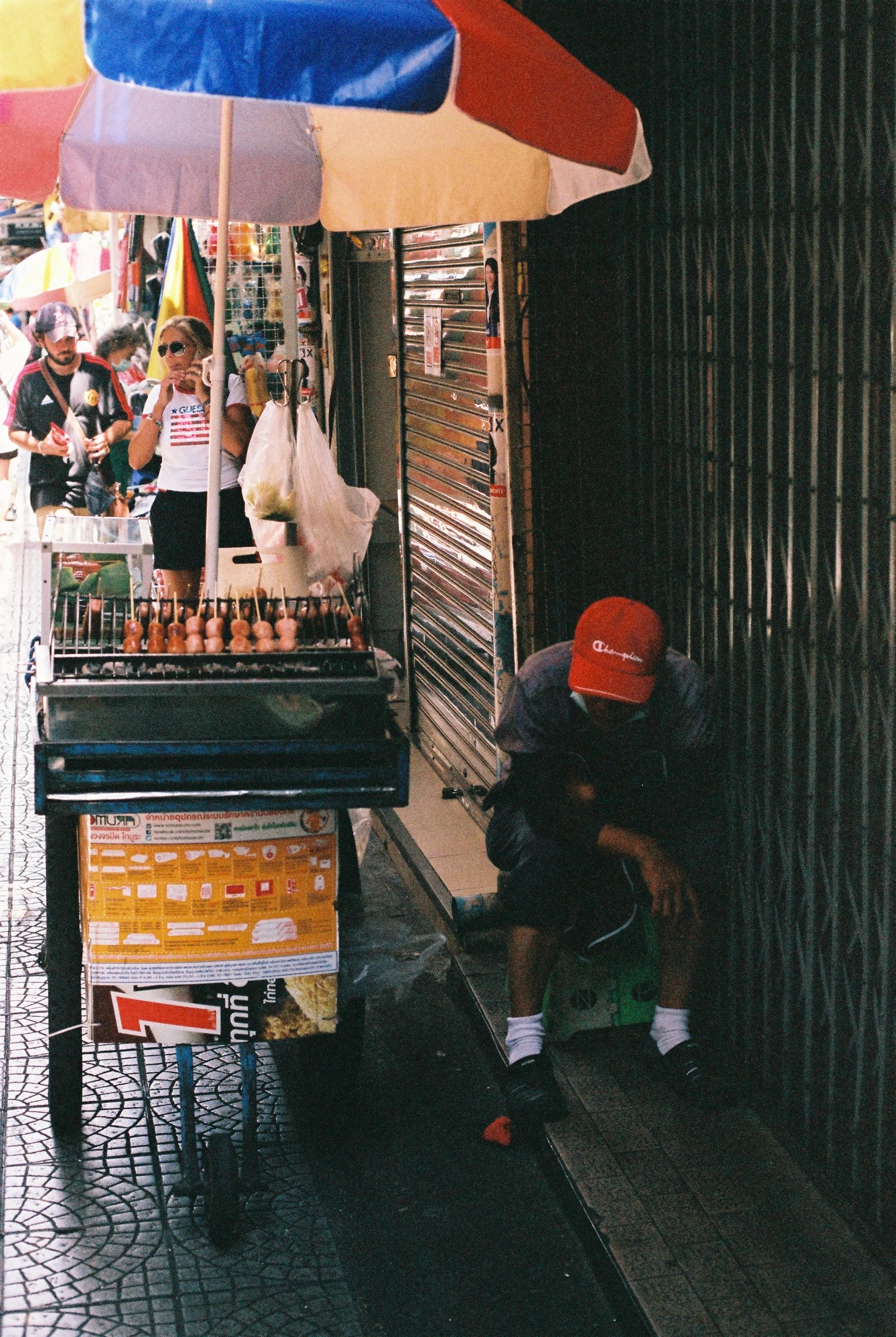 A street vendor selling grilled meat skewers under a multicolored umbrella, with a person in a red cap sitting next to the stall on a small stool in an urban area.