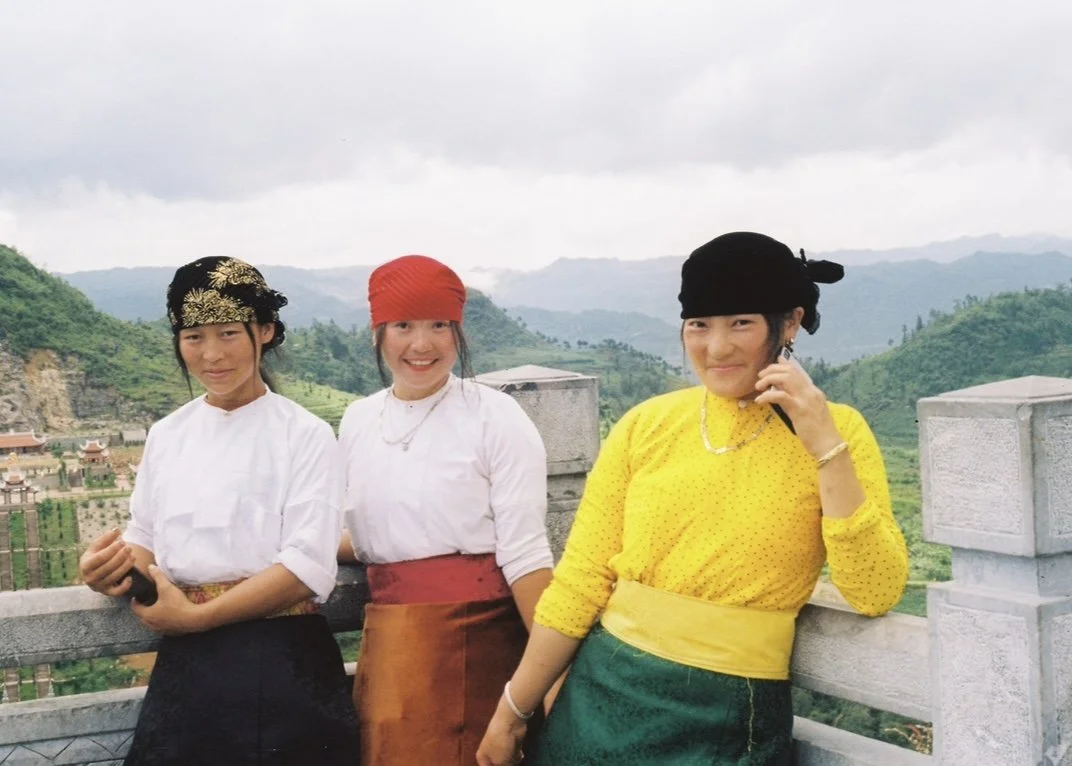 Three women dressed in traditional Asian attire, standing on a balcony with a lush, mountainous landscape in the background.