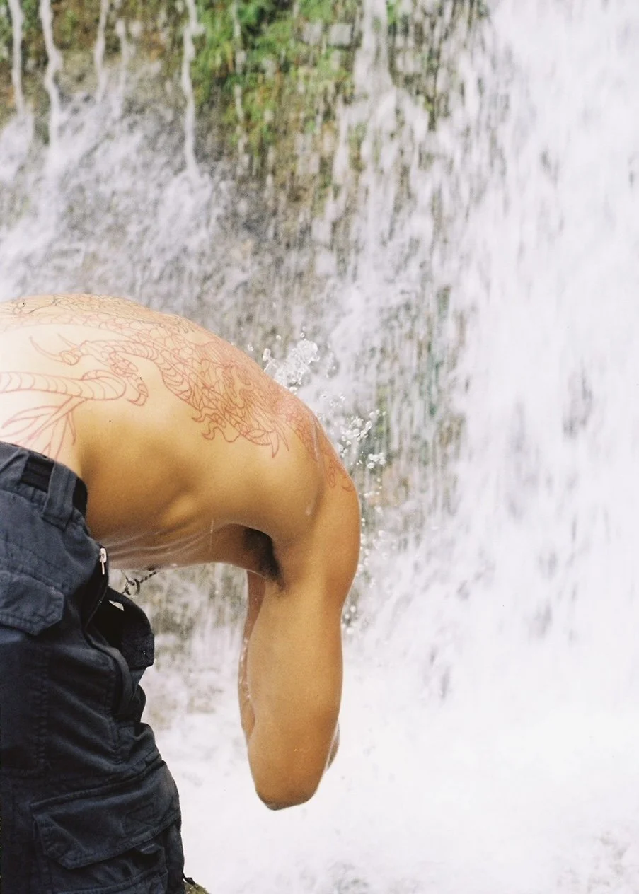 Person bending over near a waterfall with visible tattoo of a dragon on their upper back.