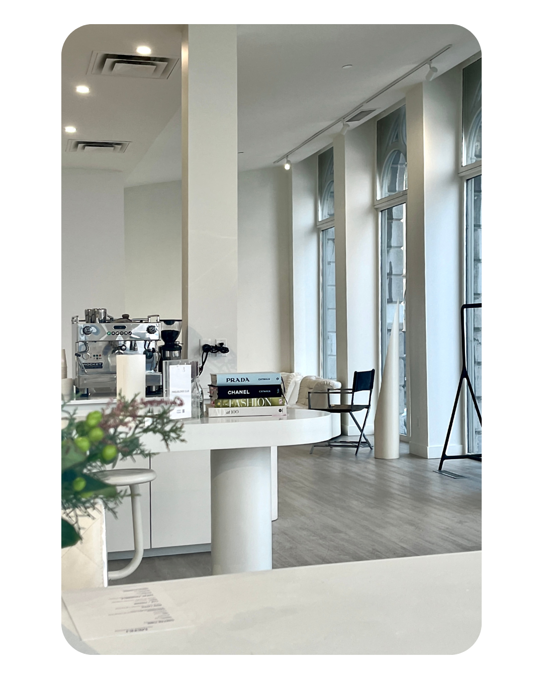 Modern interior space with large windows, white walls, and minimal furnishings including black chairs, books, and a coffee machine.