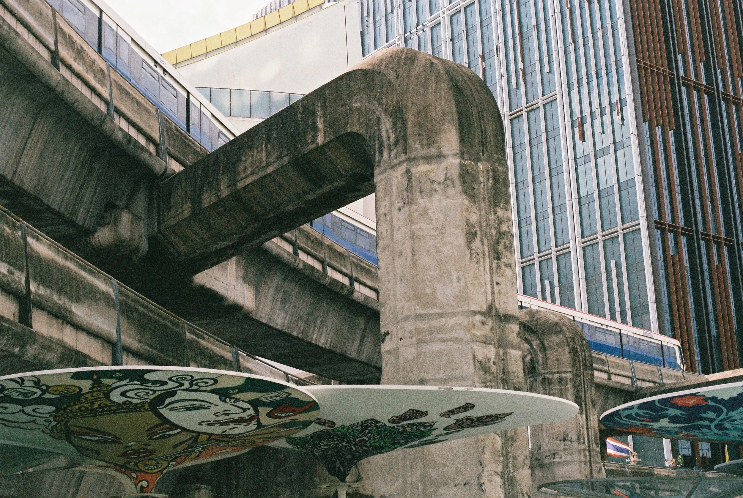 Urban scene with concrete structures and train tracks above ground, backdrop of modern glass buildings, and colorful skateboard decks suspended in the foreground.