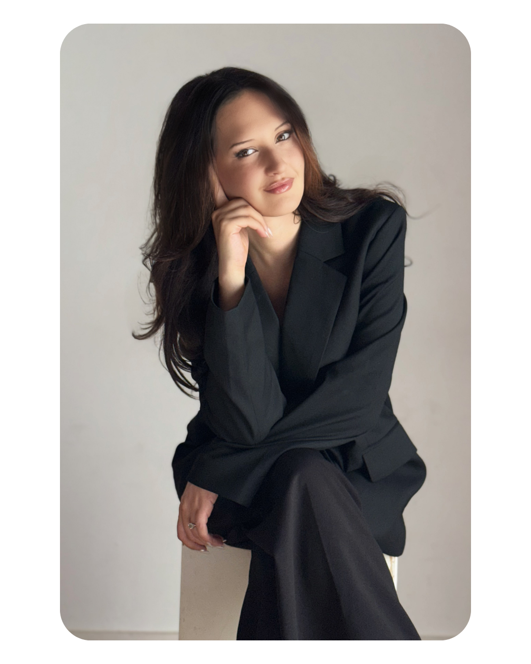 A woman with long dark hair in a black suit sitting and posing with her chin resting on her hand against a plain light-colored background.