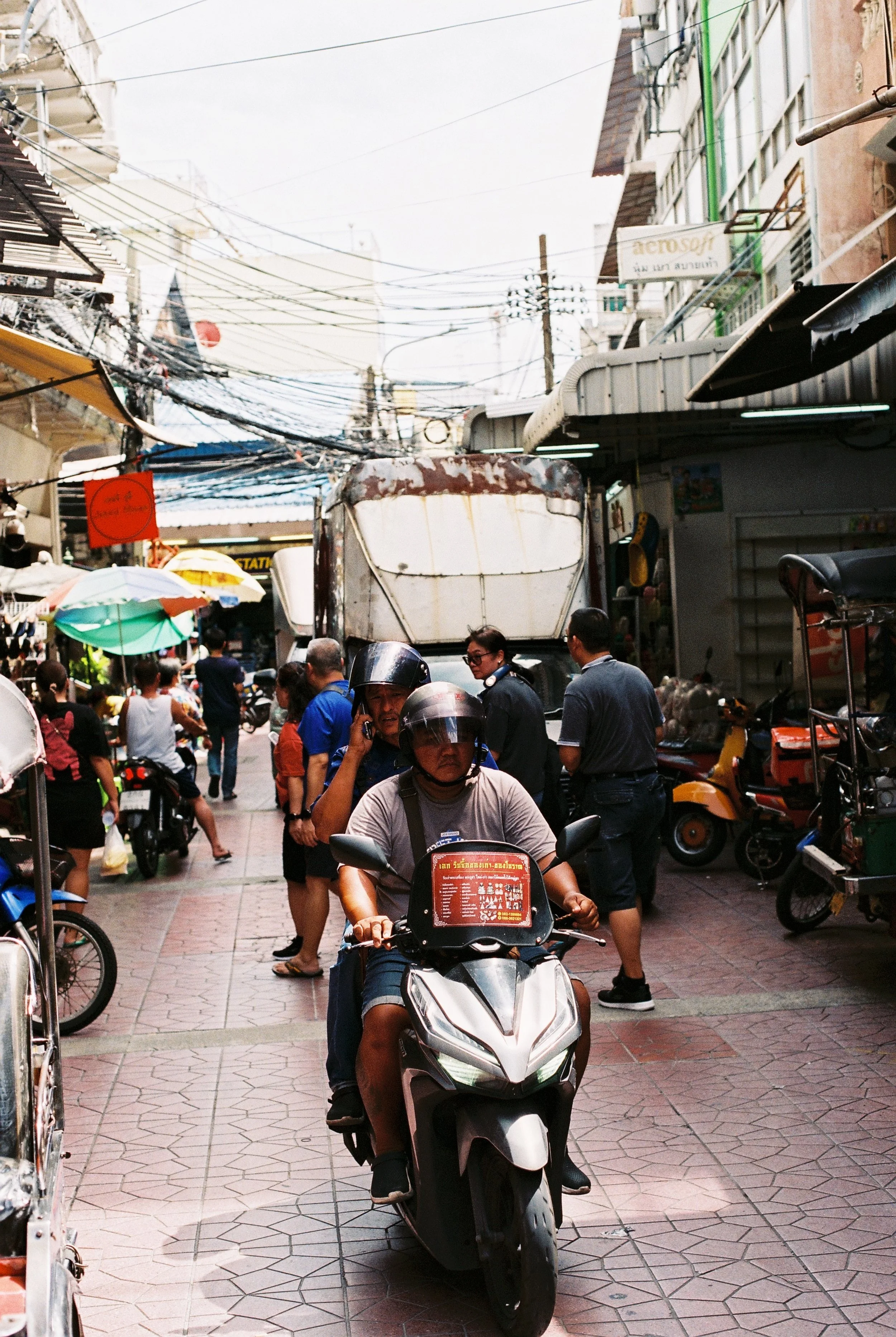 A busy street market scene in an Asian city with people walking, shopping, and riding motorcycles, colorful umbrellas and shop signage visible.