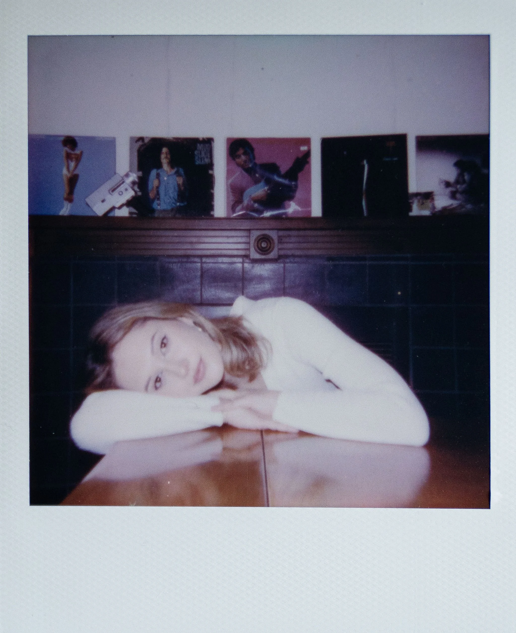 A woman with light brown hair lying on a table with her head resting on her crossed arms, looking at the camera with a neutral expression. Behind her is a dark wall with framed photos or artwork above it.