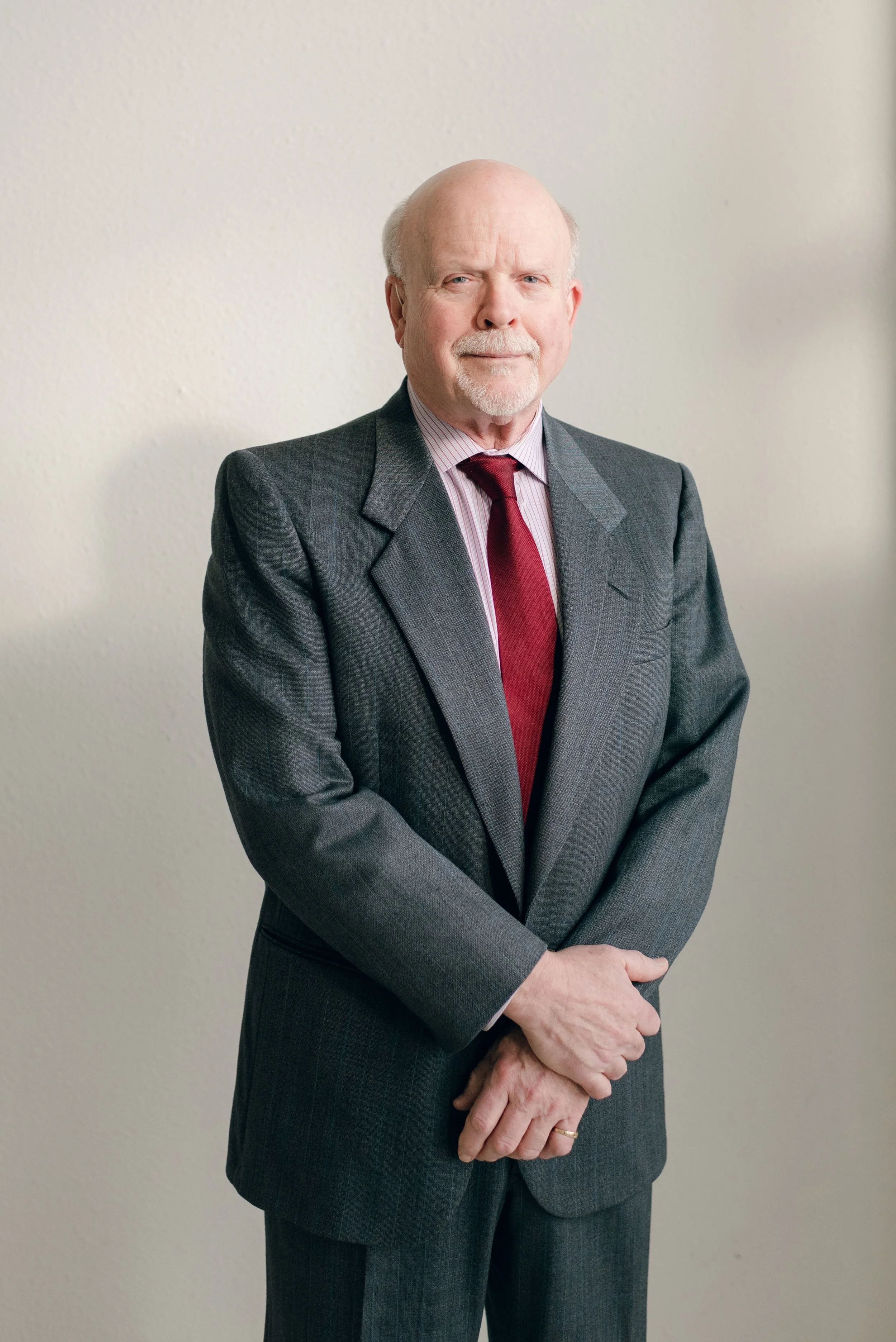 Portrait of an elderly man with a mustache and beard, wearing a dark gray suit, a pink collared shirt, and a red tie, standing against a plain off-white wall.