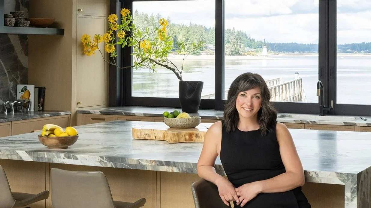 A woman sitting at a kitchen island with a large window behind her showing a body of water and a lighthouse in the distance. The kitchen has a modern design with a marble countertop, a bowl of lemons, and a decorative branch with yellow flowers in a black vase in front of the window.