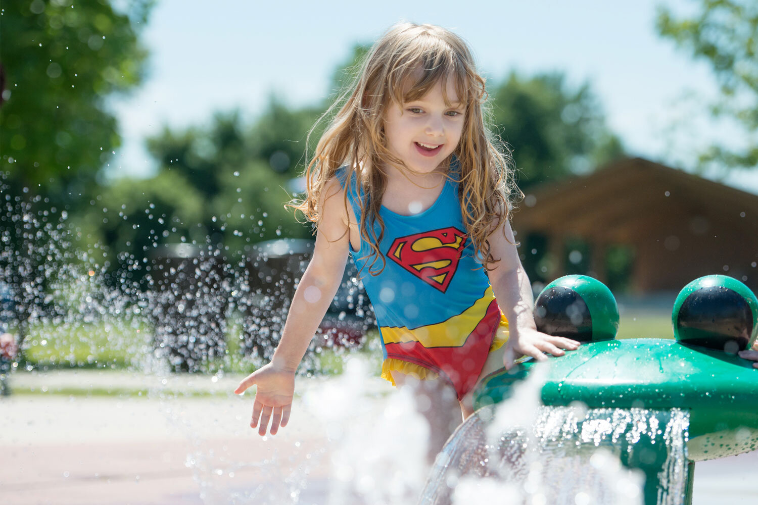 little girl in superman bathing suit playing in a water fountain